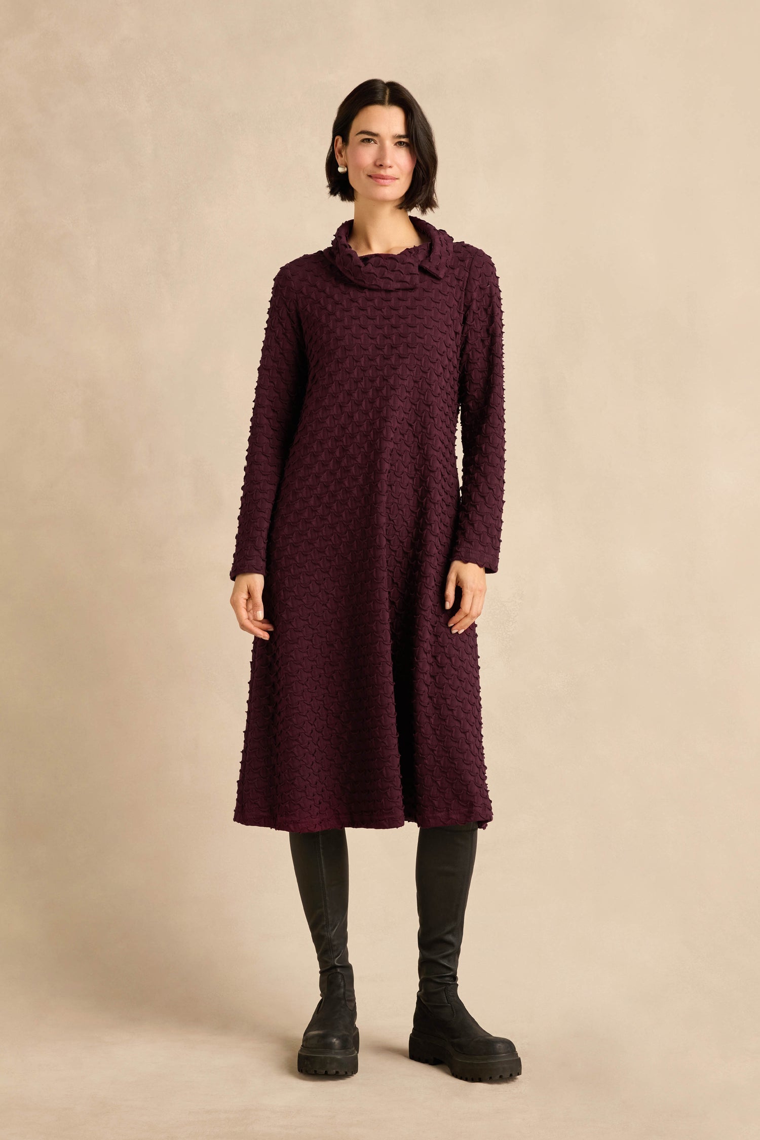A person stands against a beige background wearing the Origami Pocket Jersey Cowl Neck Dress—a textured, dark purple fit-and-flare dress—paired with black leggings and black boots.