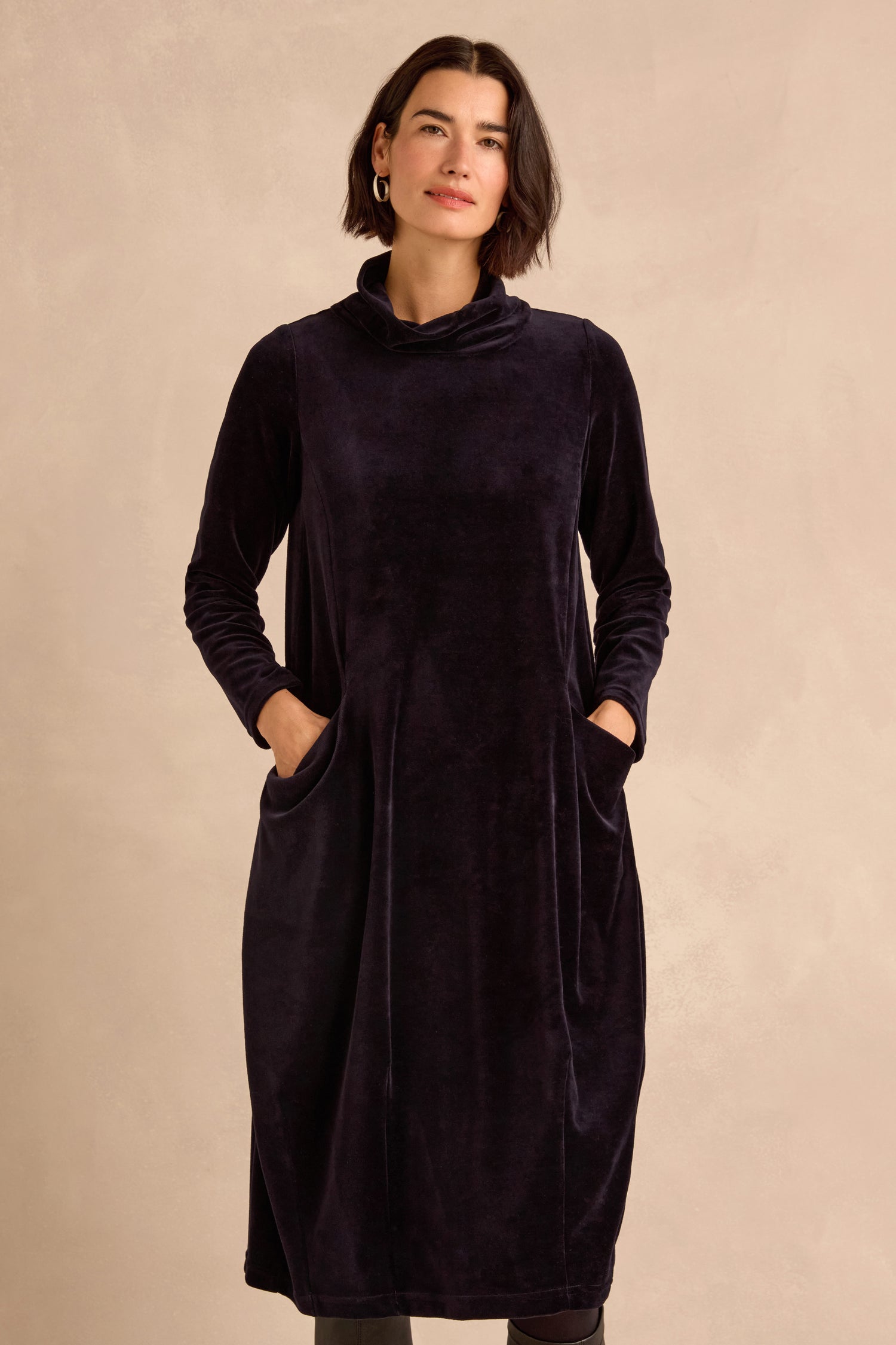A woman models the Velvet Jersey Pocket Dress—a long-sleeved, black velvet jersey dress featuring pockets and a subtle cowl neck—while standing against a neutral beige background.