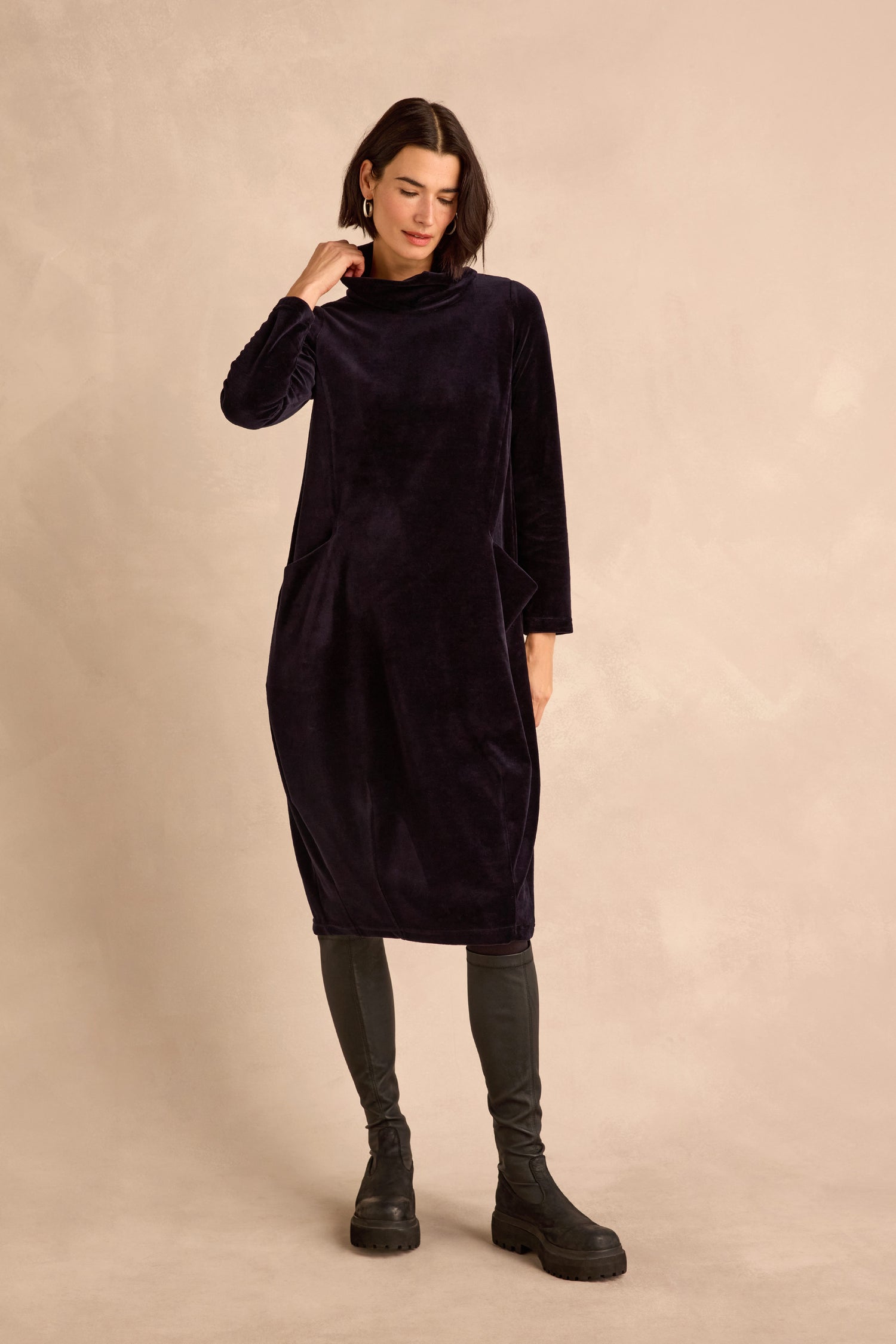 A woman wearing the Velvet Jersey Pocket Dress—a long-sleeve, dark velvet jersey dress featuring a cowl neck and side pockets—paired with dark leggings and black boots, stands against a neutral beige background.