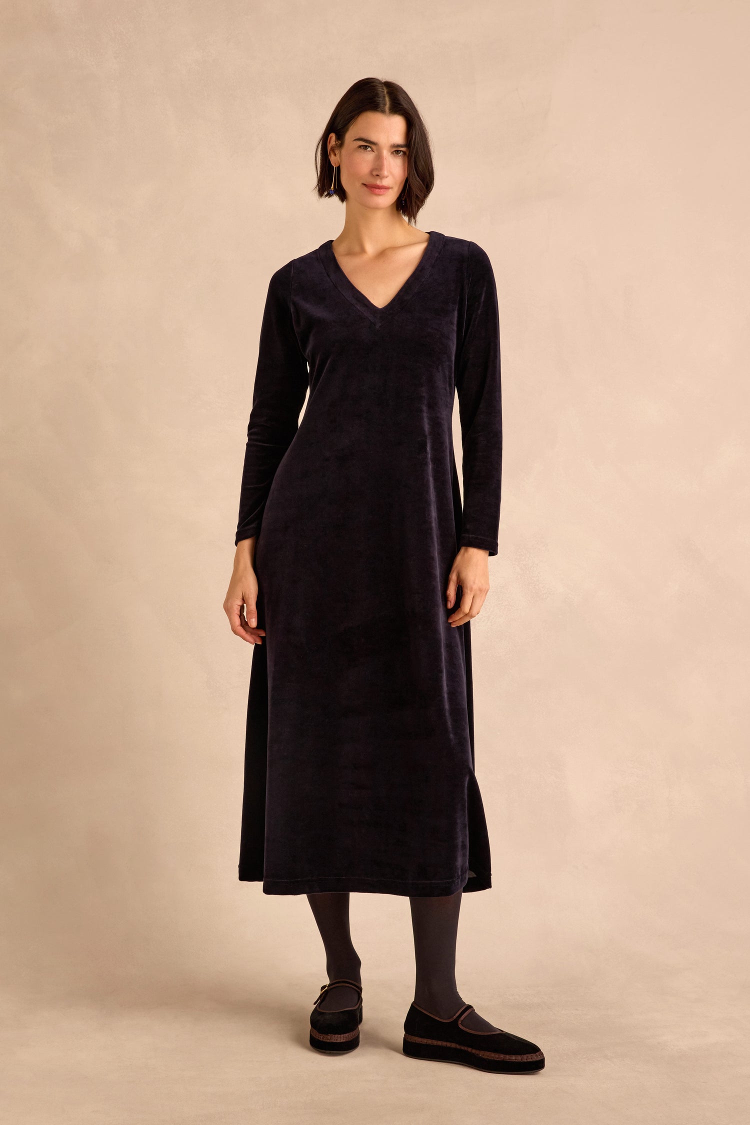 A woman stands against a beige background wearing the Night Velvet Jersey Maxi Dress—a long-sleeve, cotton-rich velvet maxi dress with a V-neckline—paired with dark tights and flat shoes.