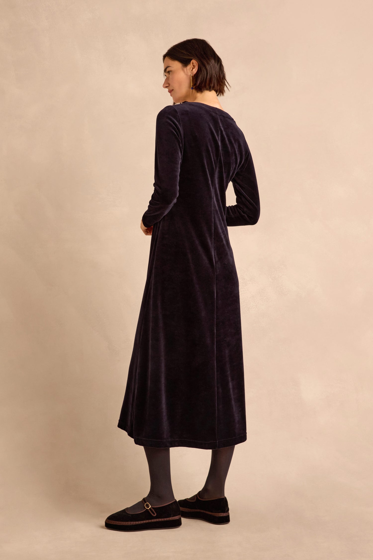 A woman stands sideways against a light beige background, wearing the Night Velvet Jersey Maxi Dress with black tights and black shoes.
