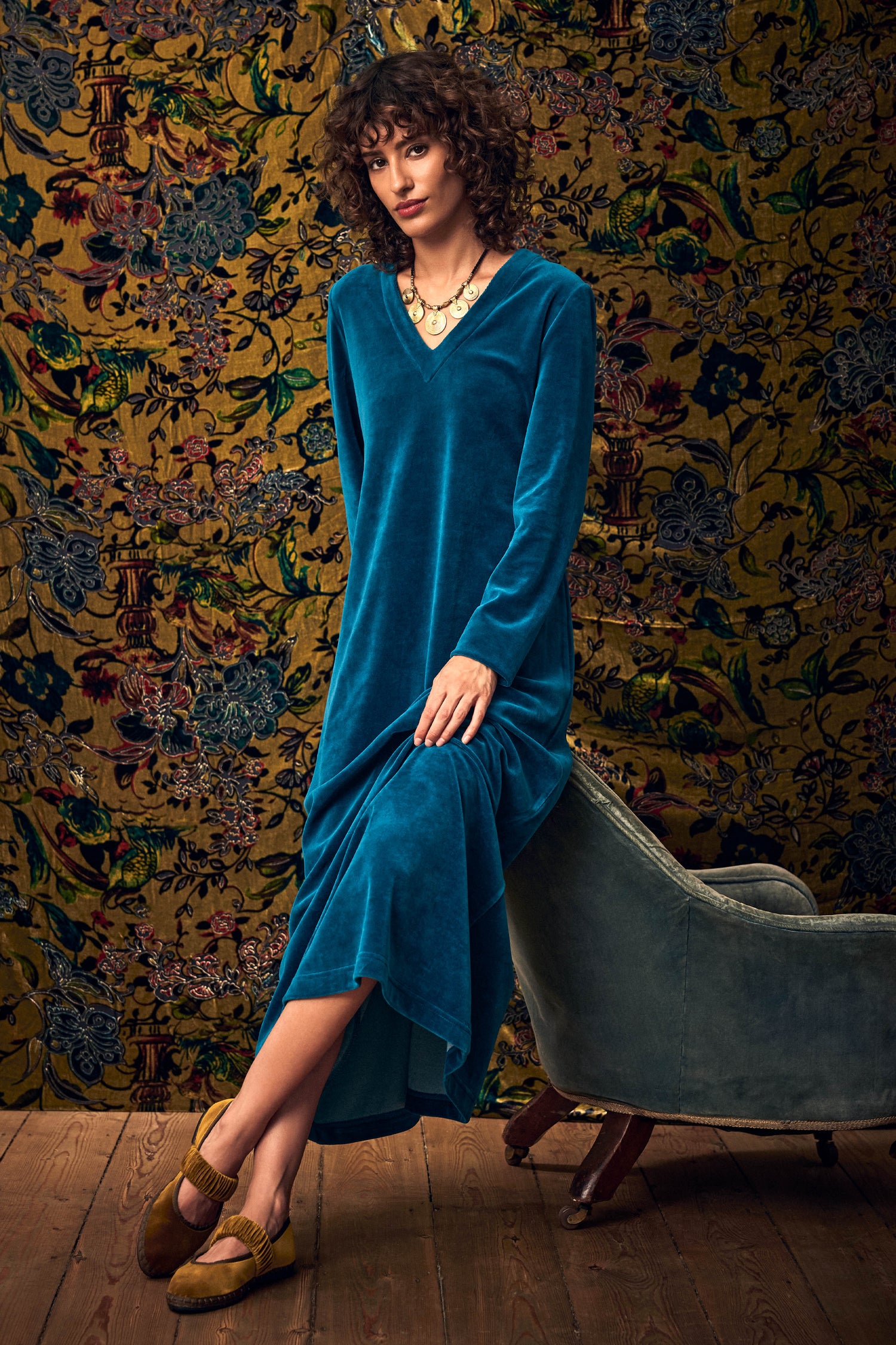 A woman in a teal Teal Velvet Jersey Maxi Dress and yellow flat shoes leans against a chair.