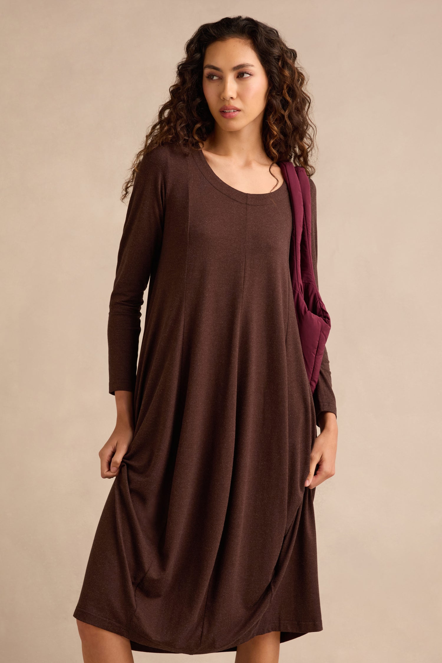 A woman stands against a beige background wearing the Soft Viscose Jersey Bubble Dress in brown with long sleeves, carrying a maroon bag over her shoulder.