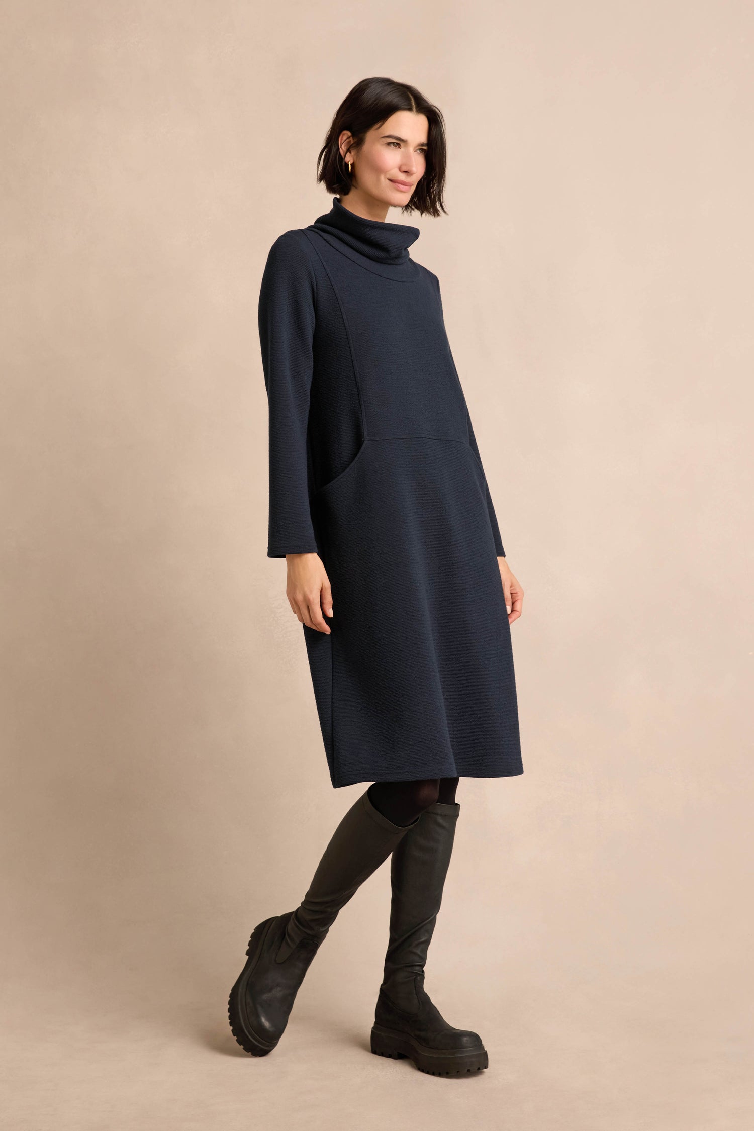 Woman wearing a long-sleeve, navy blue Double Jersey Crinkle Dress with black leggings and chunky black boots, standing against a plain beige background.