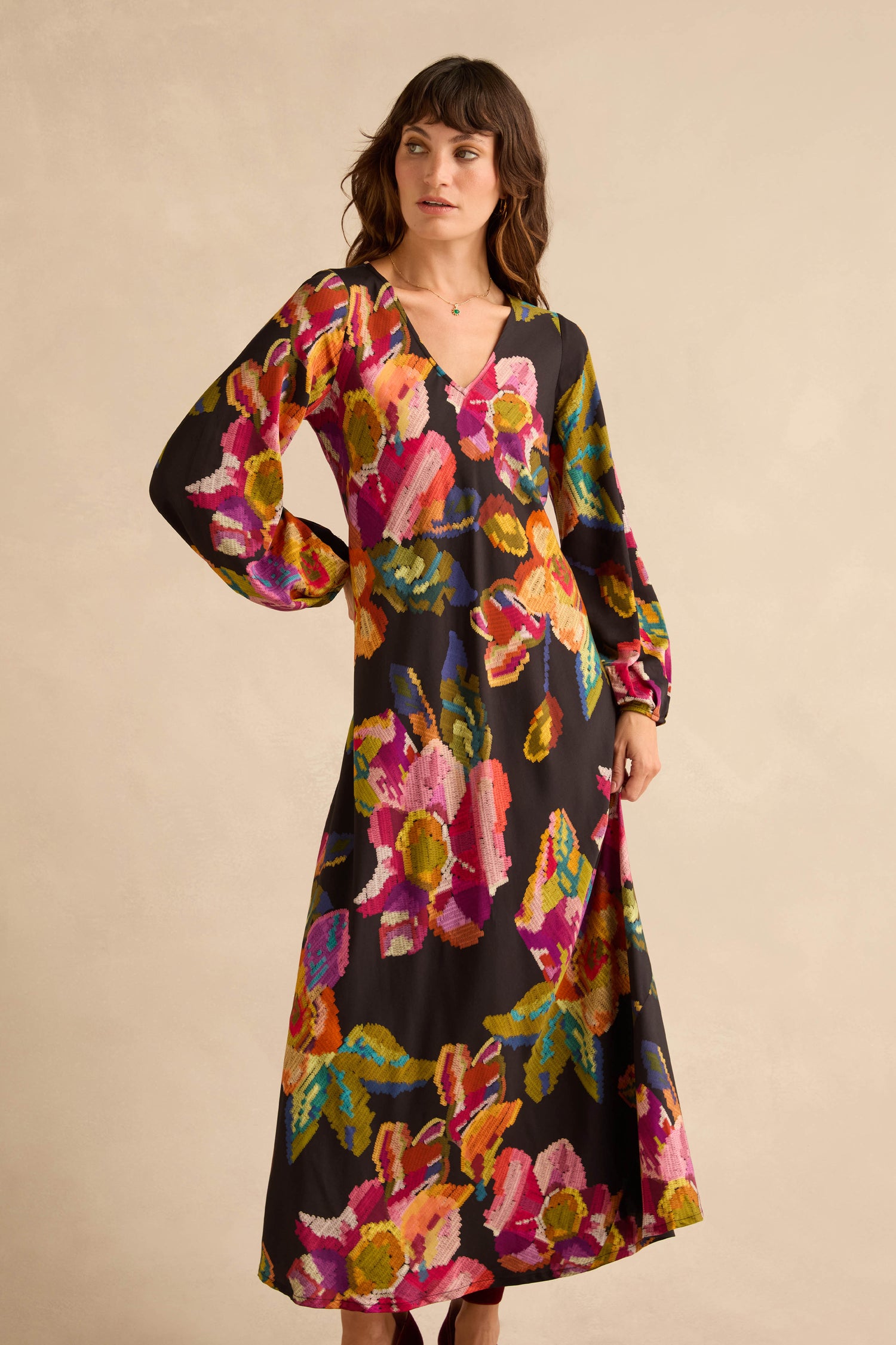 A woman wears a long-sleeved, v-neck Pixelated Floral Flared Dress in pure viscose, featuring a black background with a multicolored pattern.