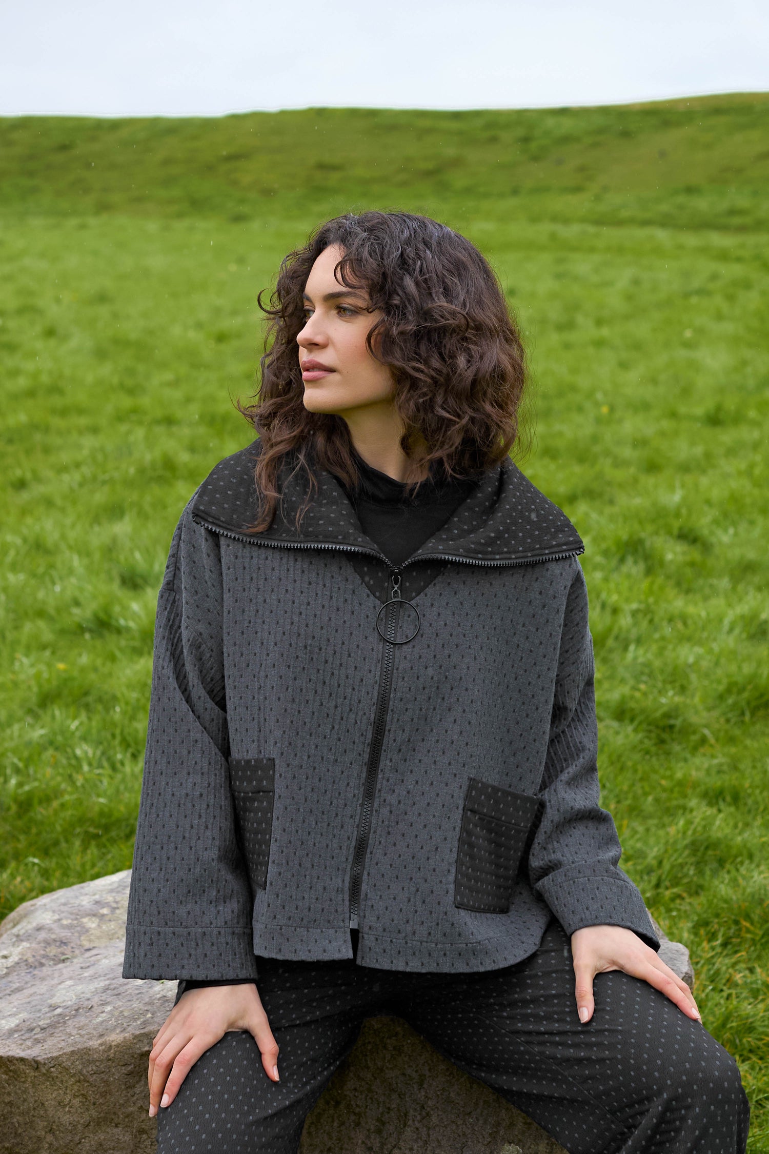 A person with curly hair sits on a rock in a grassy field, wearing the Square Jacquard Boxy Jacket along with coordinating dark pants.