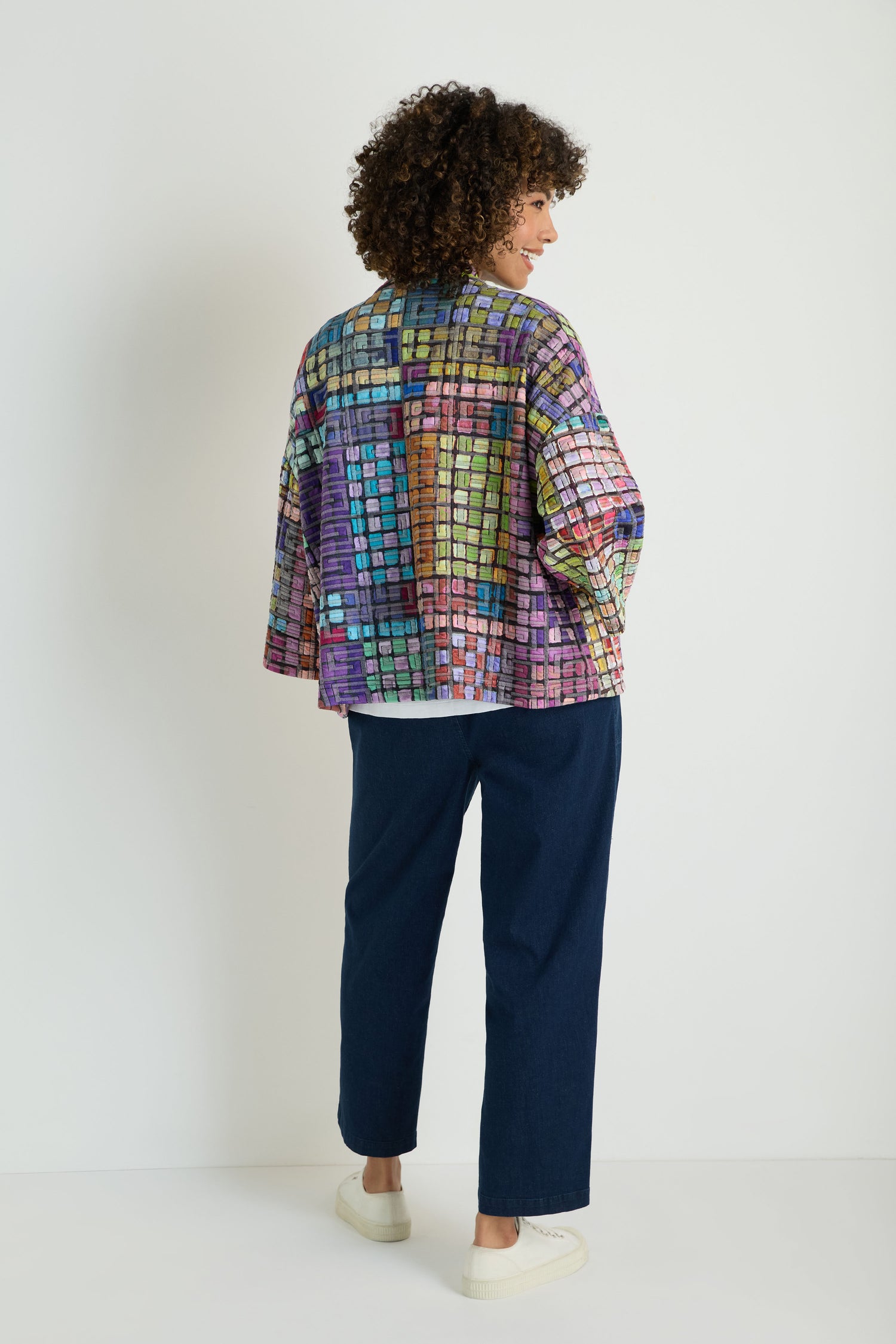 A person wears the Coloured Glass Tiles Jersey Jacket, featuring a multicolored grid design like colored glass tiles, over a blue outfit; their hand is tucked into one of the jacket’s front pockets.