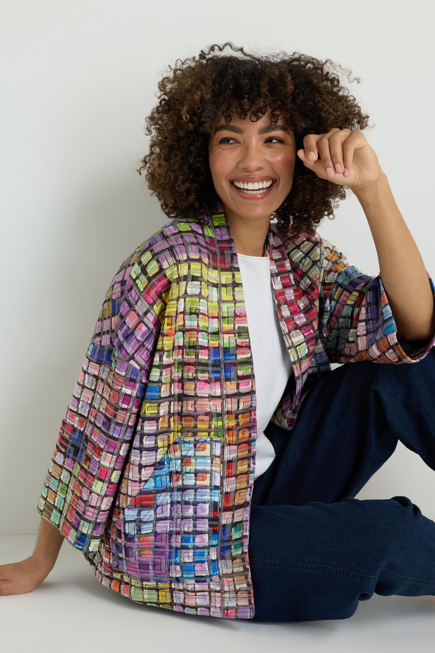 A woman wears the Coloured Glass Tiles Jersey Jacket—a lightweight blue top and pants set with vibrant glass tile patterns—paired with white sneakers against a plain light background.