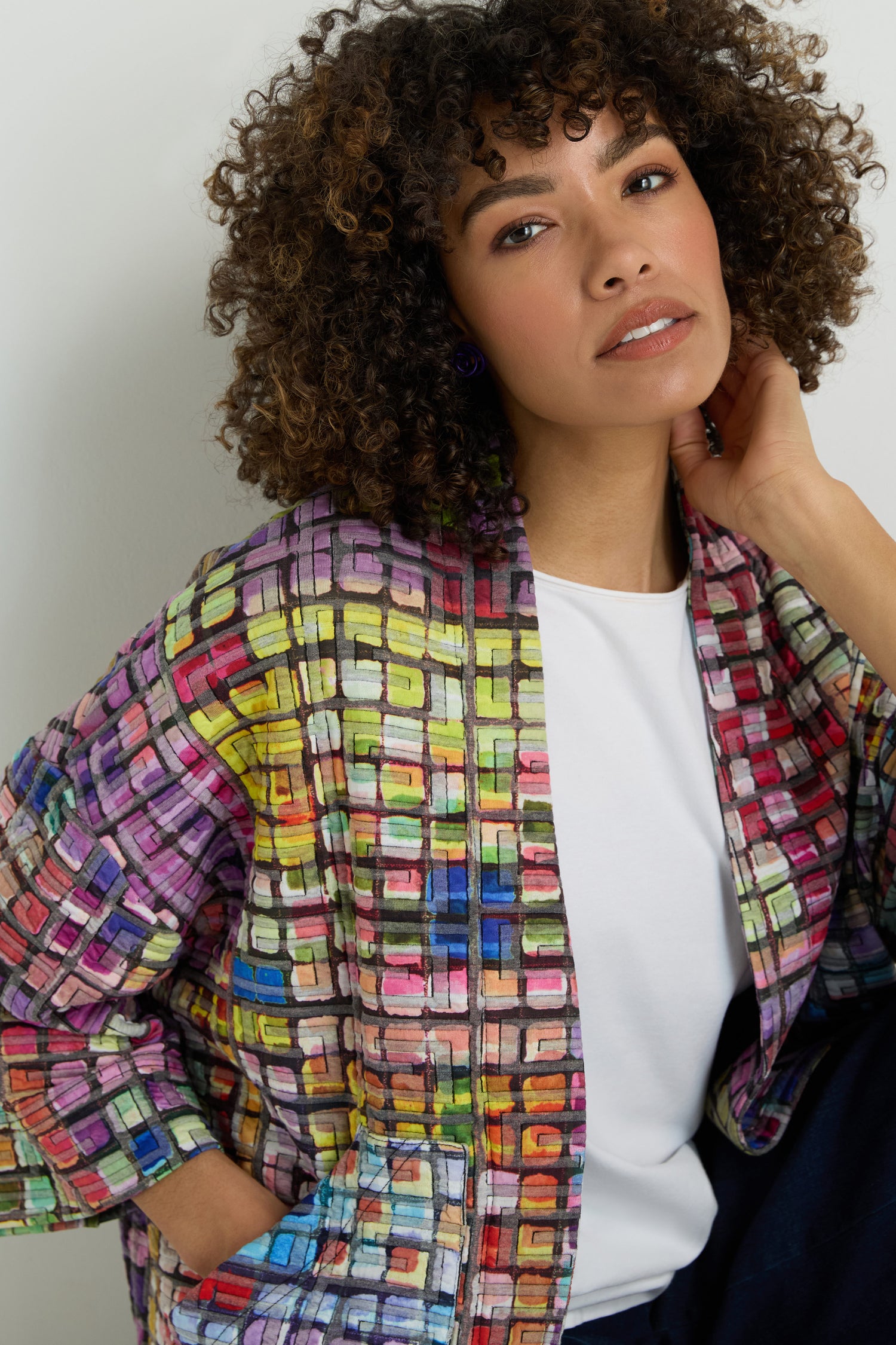 A woman models the Coloured Glass Tiles Jersey Jacket, featuring a vibrant mosaic pattern, over a blue top and blue pants against a plain light background.