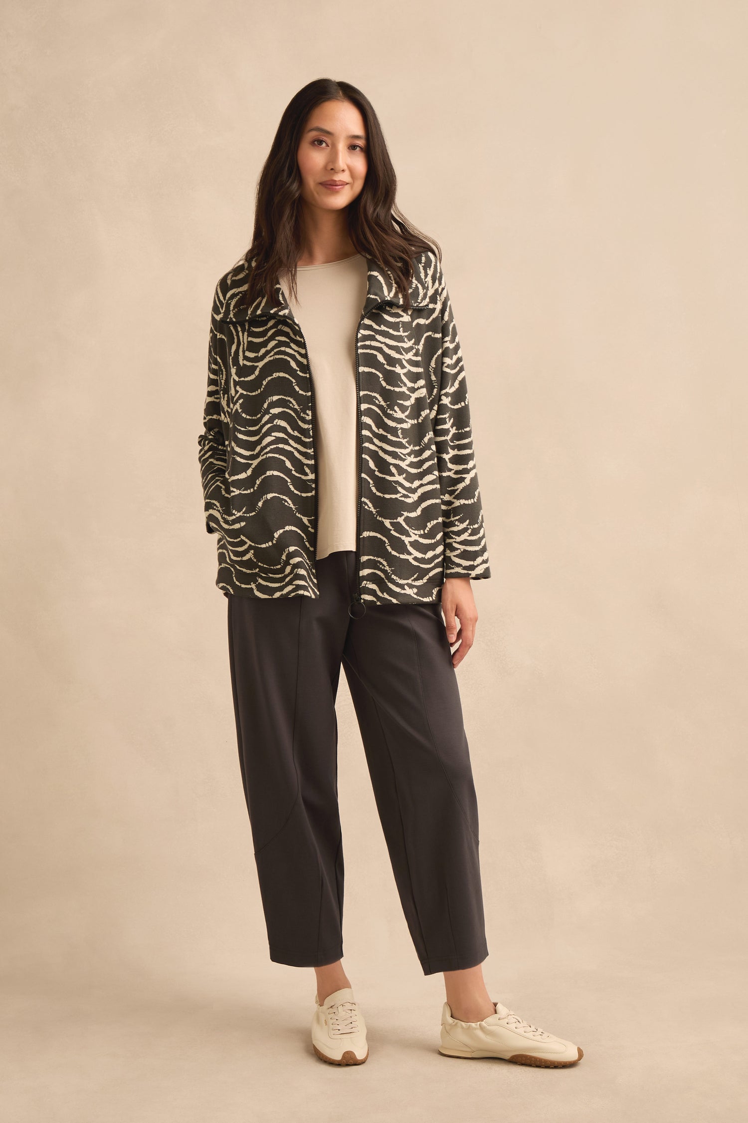 A woman stands against a beige background wearing the Dancing Waves Zip Boxy Jacket—a black and white wave motif jersey piece—paired with a cream top, dark cropped pants, and white sneakers.