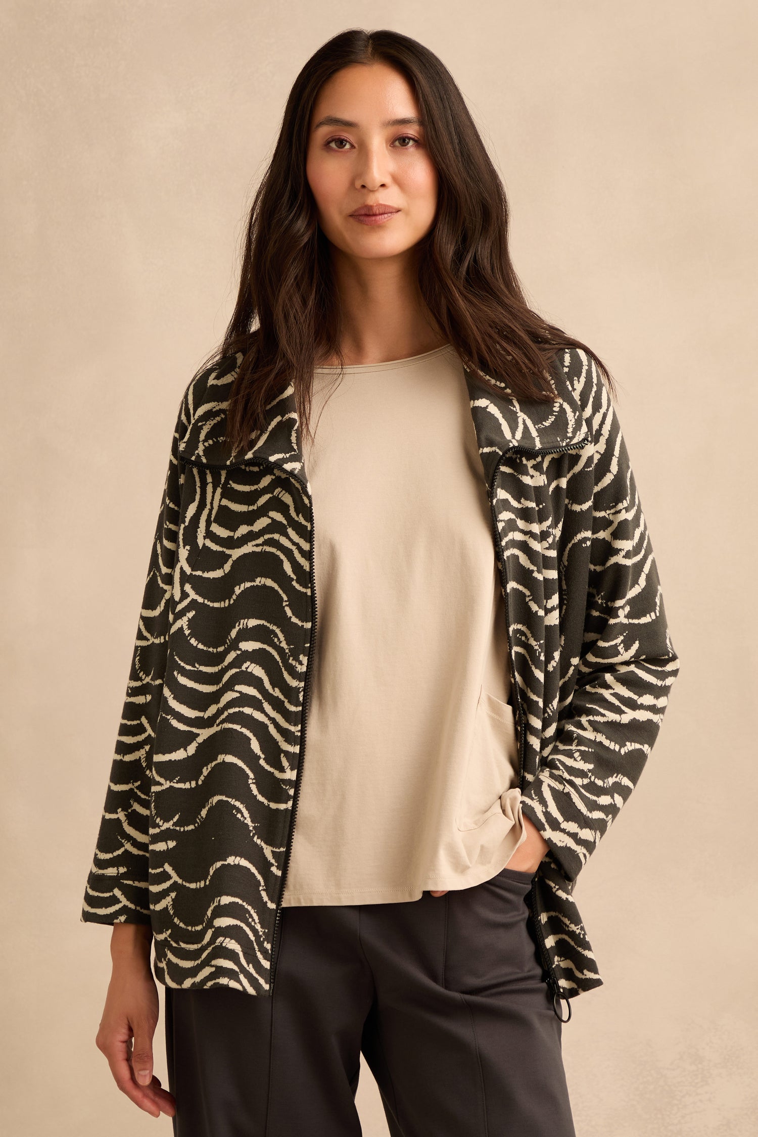 A woman stands against a neutral background, wearing the Dancing Waves Zip Boxy Jacket—a black and white wave motif jersey piece—layered over a light shirt and dark pants, with one hand in her pocket.