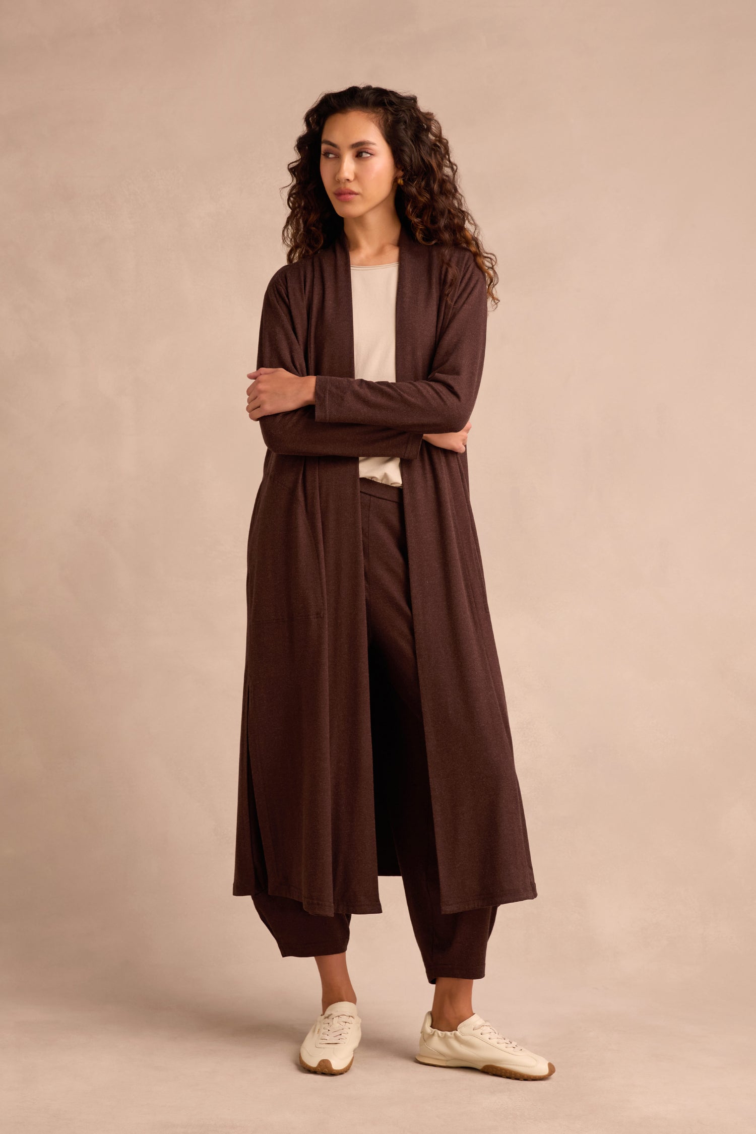 A woman stands with her arms crossed, wearing the Soft Viscose Jersey Long Line Cardi in soft brown, paired with matching pants, a cream shirt, and white sneakers against a neutral background.