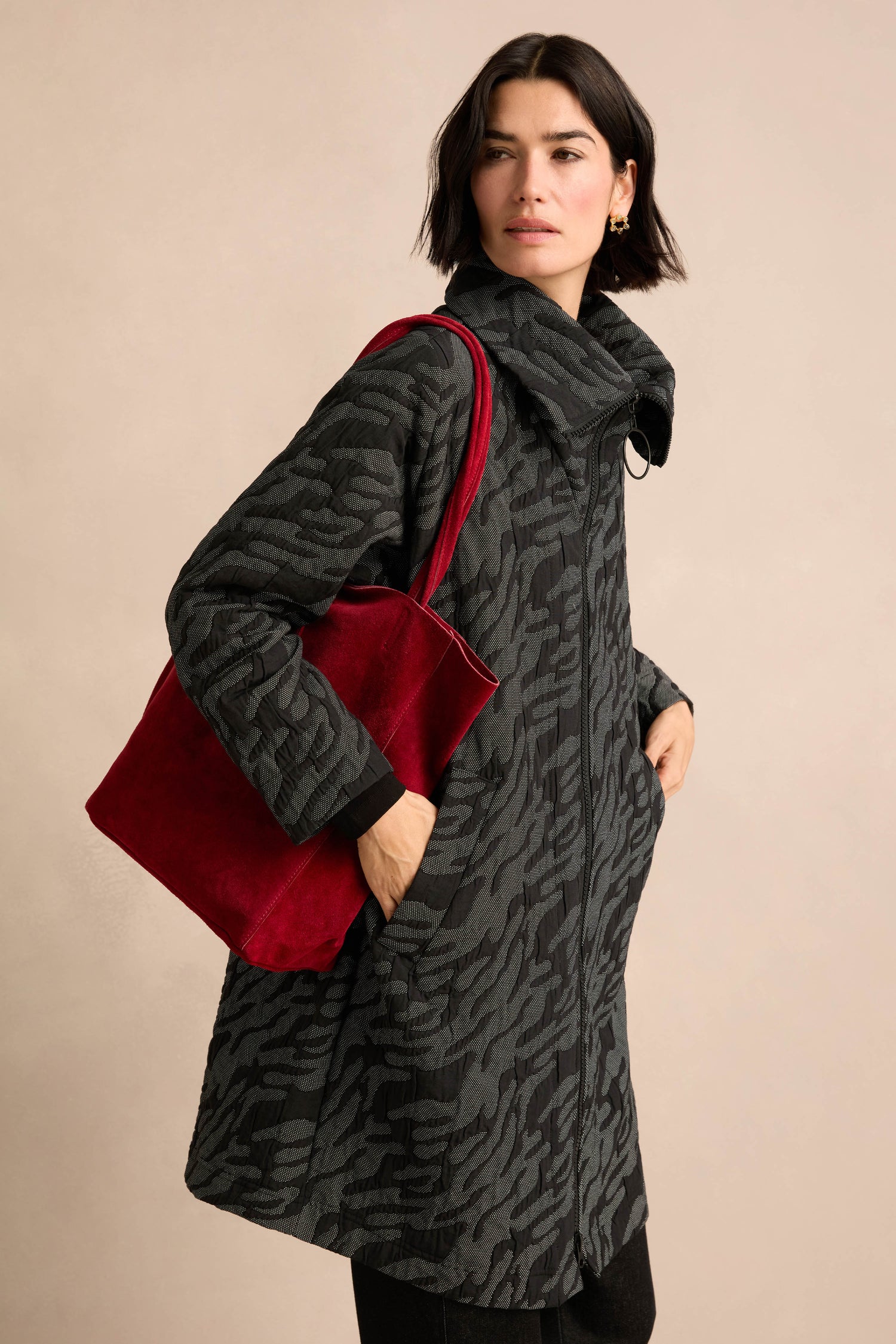 A woman models the Quilted Coat with an oversized silhouette and bold pattern, holding a large red handbag over her shoulder against a neutral background.