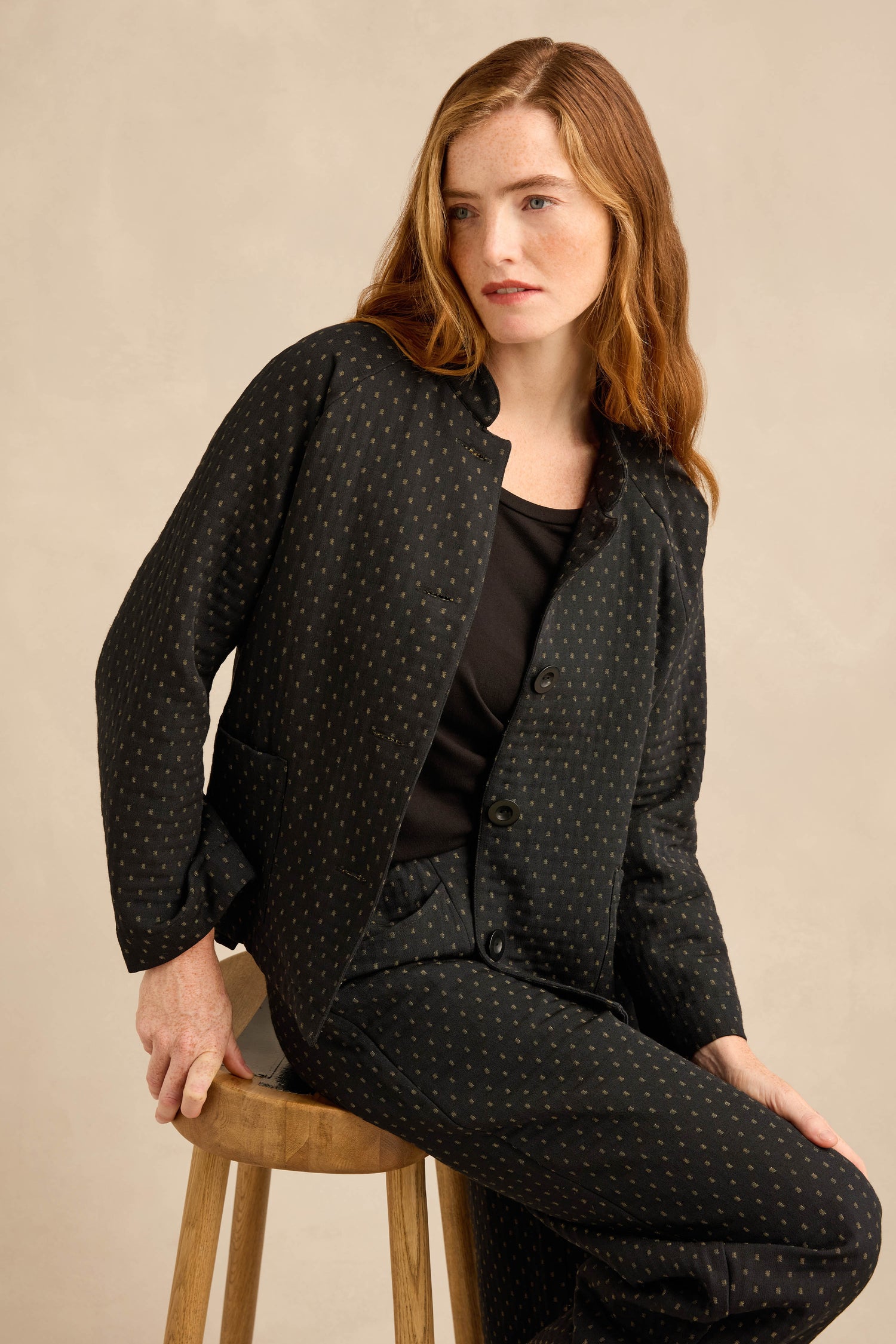 A woman sits on a wooden stool wearing a workwear-inspired Square Jacquard Jacket with matching trousers and a black top underneath.