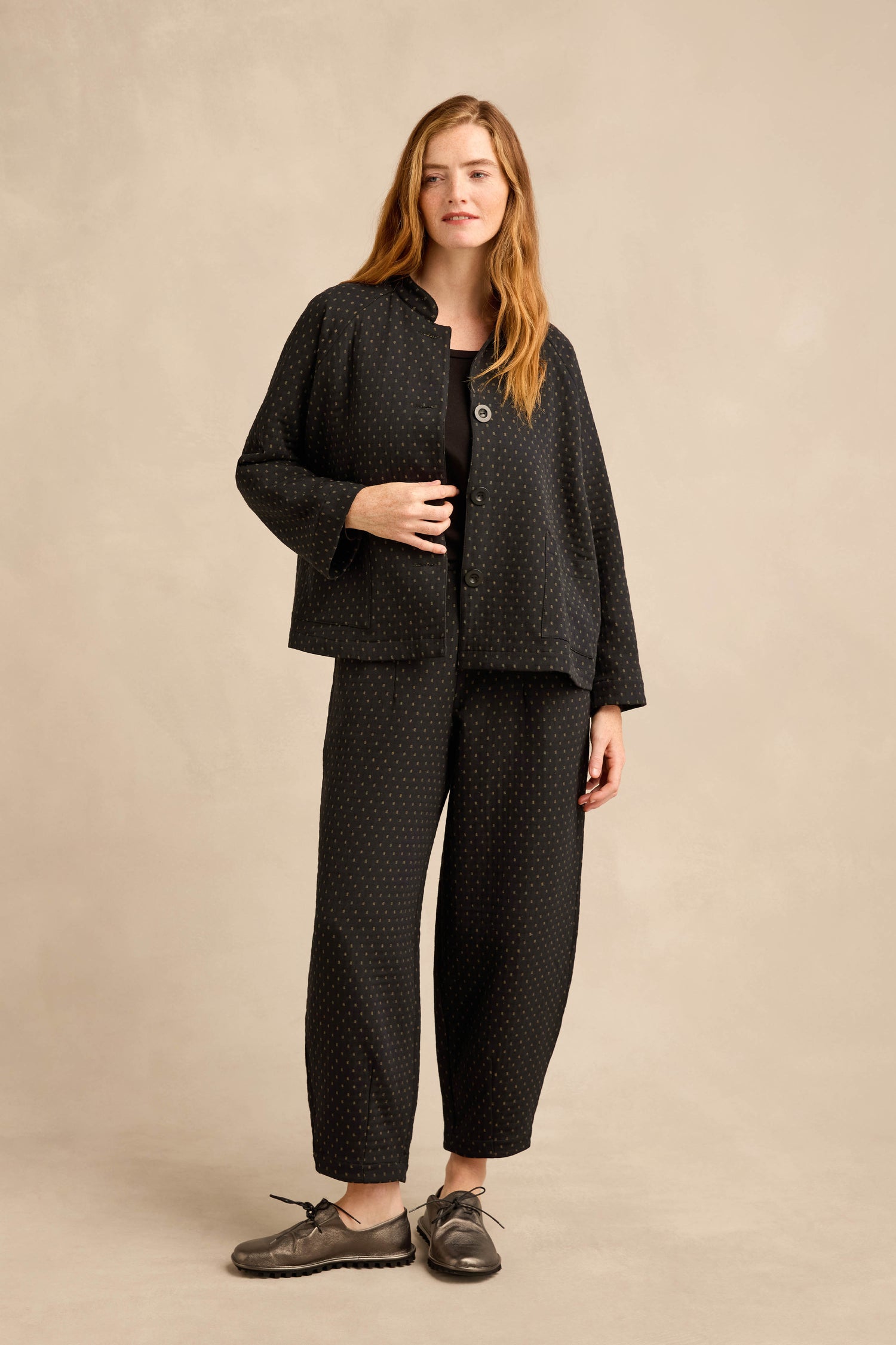 A woman stands against a beige background wearing a loose-fitting, workwear-inspired Square Jacquard Jacket with black polka dots, matching wide-leg pants, a black top, and gray shoes.