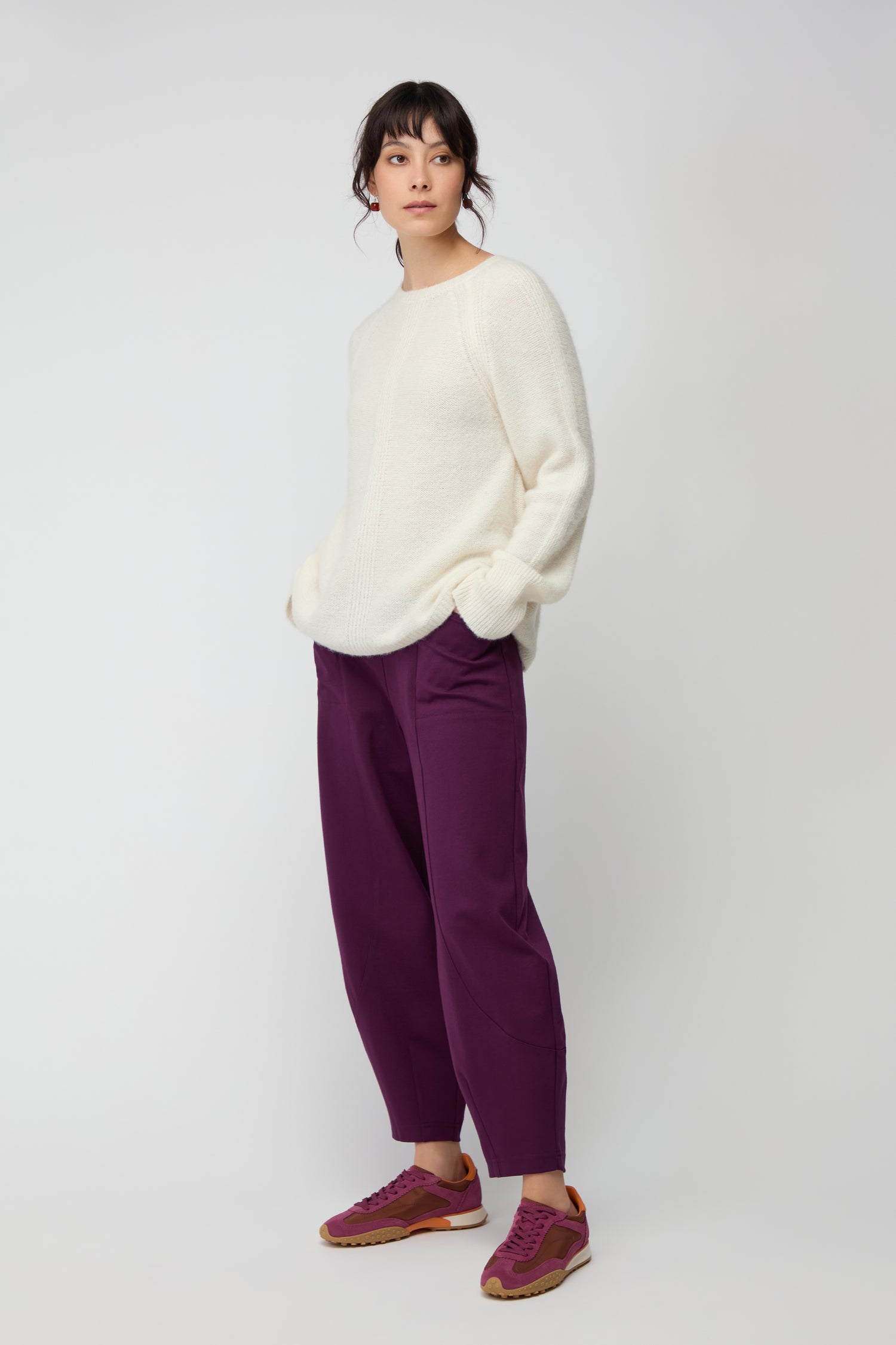 A person stands against a plain light background wearing white sweater and loose purple Ponte Bubble Trousers with an elasticated waistband, paired with purple sneakers that have orange accents.