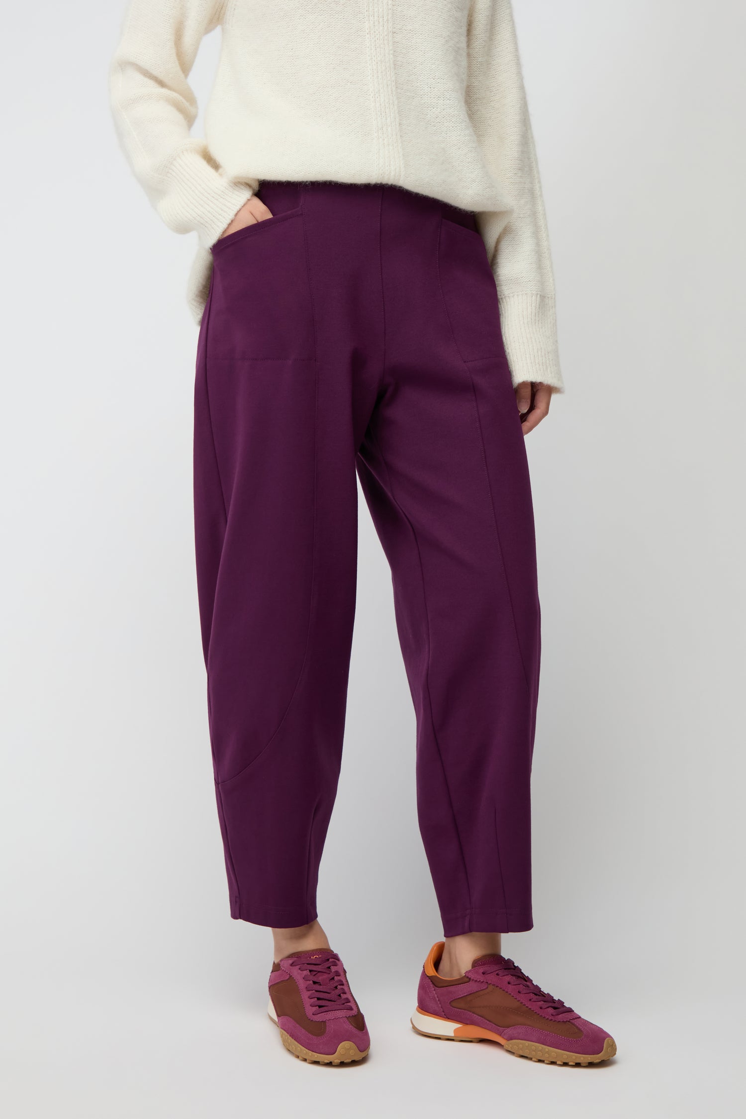 Person wearing a cream sweater, Ponte Bubble Trousers in purple—a true wardrobe staple—and purple sneakers with brown soles, standing against a plain light background.