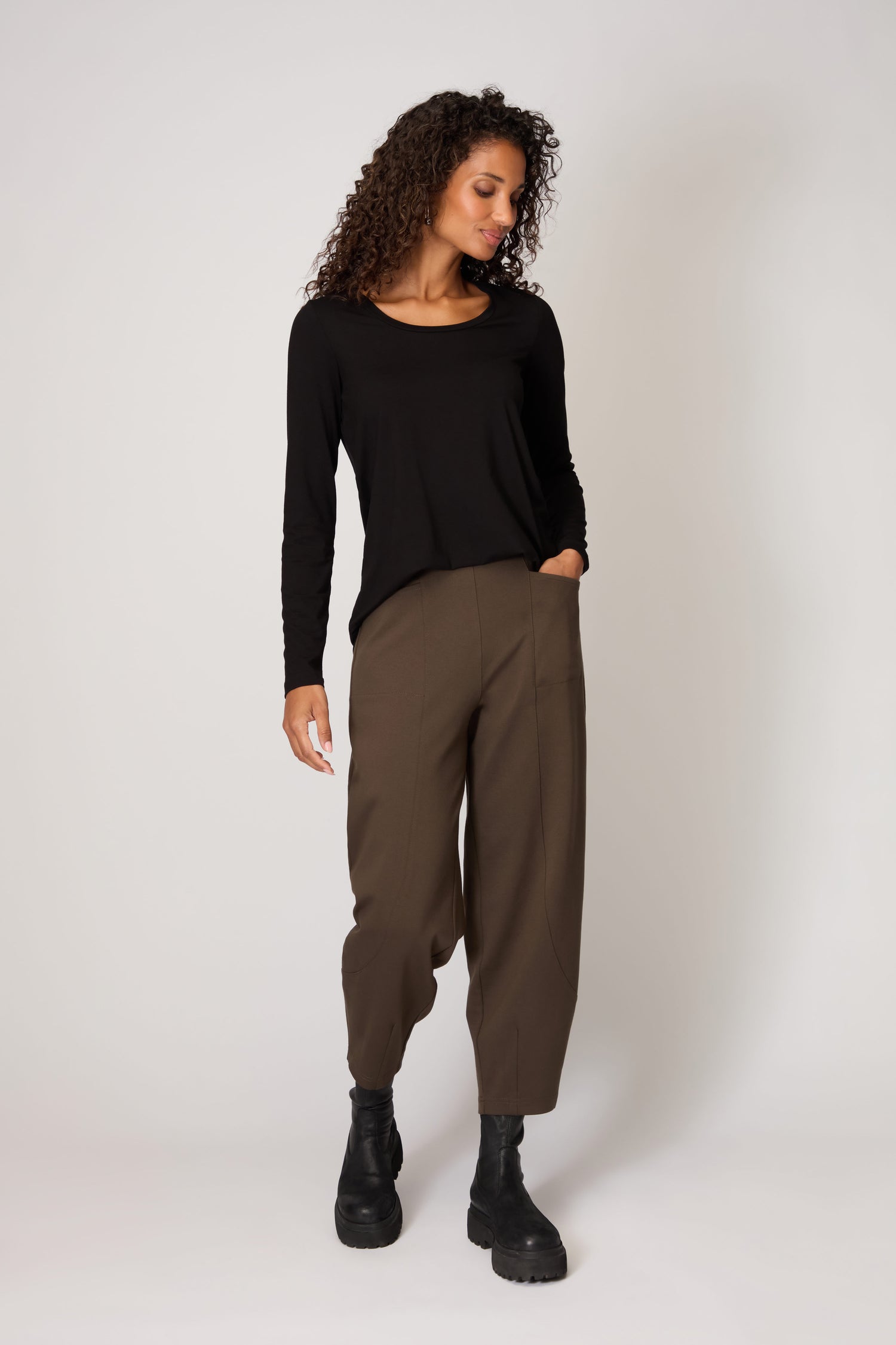 A person stands against a plain background wearing a long-sleeve black top, black boots, and Ponte Bubble Trousers with an elasticated waistband, one hand in their pocket and looking slightly downward.