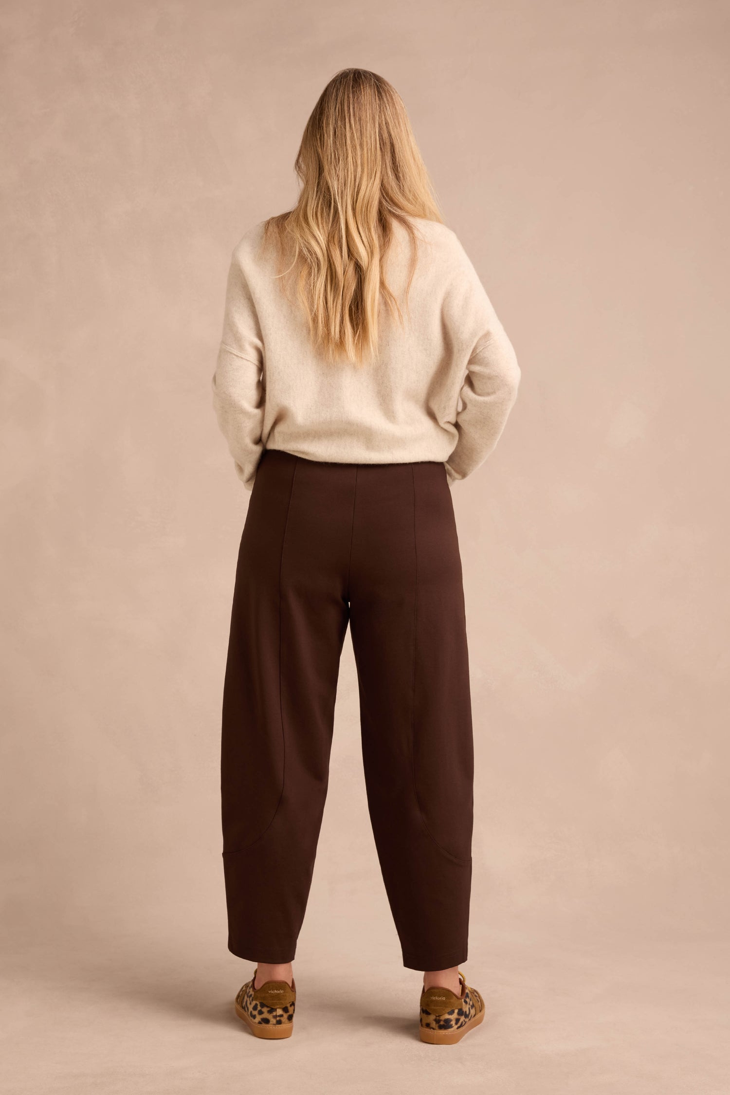 A person with long blonde hair stands facing away, wearing a beige sweater, the Chocolate Ponte Bubble Trousers with a rounded leg, and leopard print slip-on shoes against a neutral background.