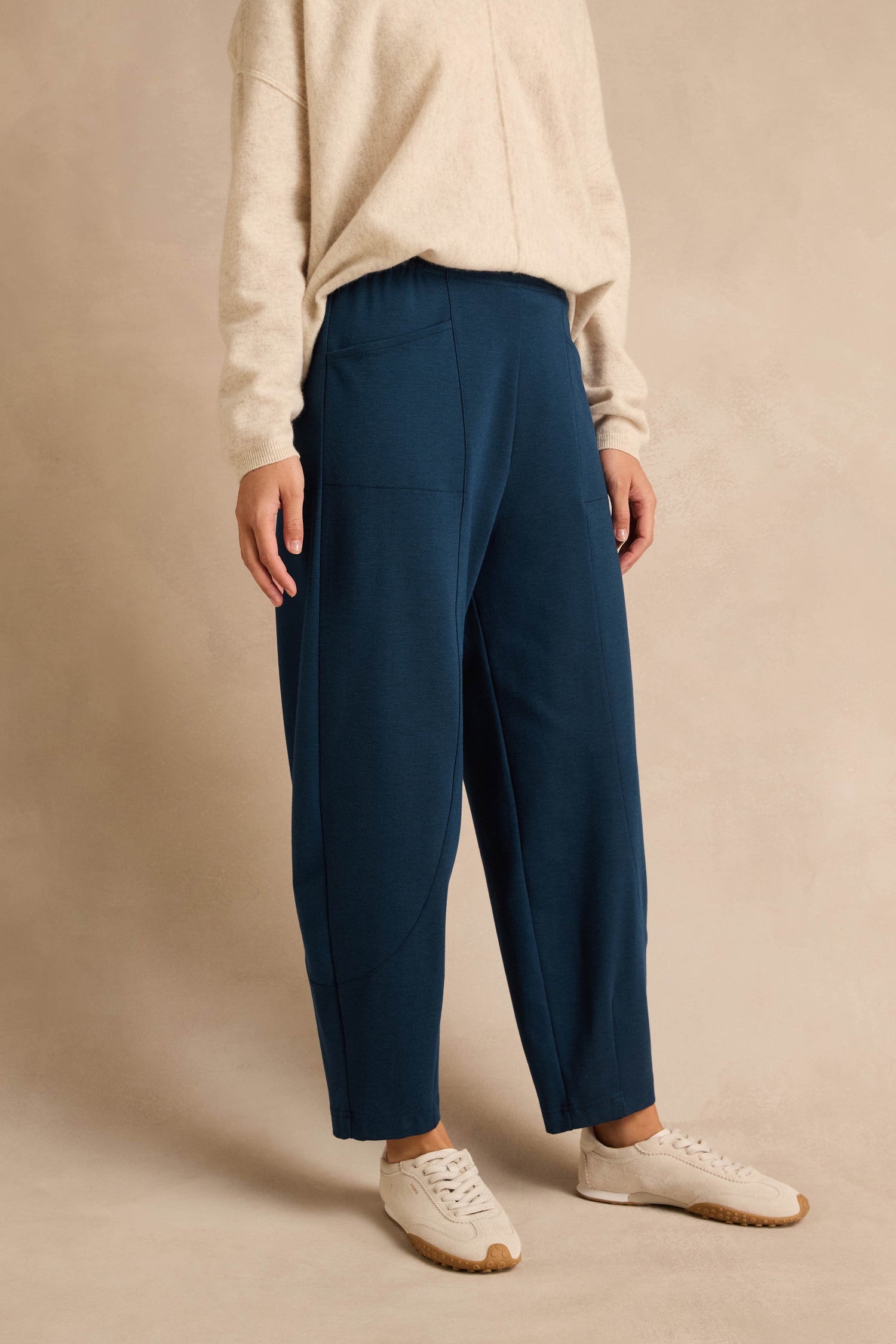 A person wearing a beige sweater, Marine Ponte Bubble Trousers with a loose, rounded leg silhouette in blue, and white sneakers stands against a plain beige background.