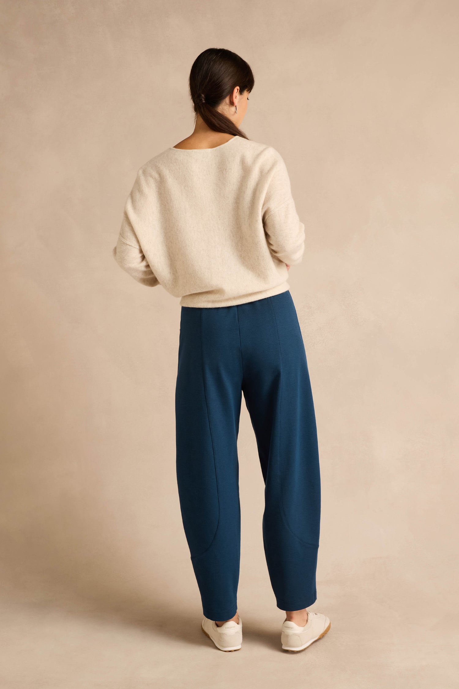 A person stands facing away, wearing a cream sweater and the Marine Ponte Bubble Trousers in blue, featuring a rounded leg silhouette, paired with white shoes against a plain beige background.