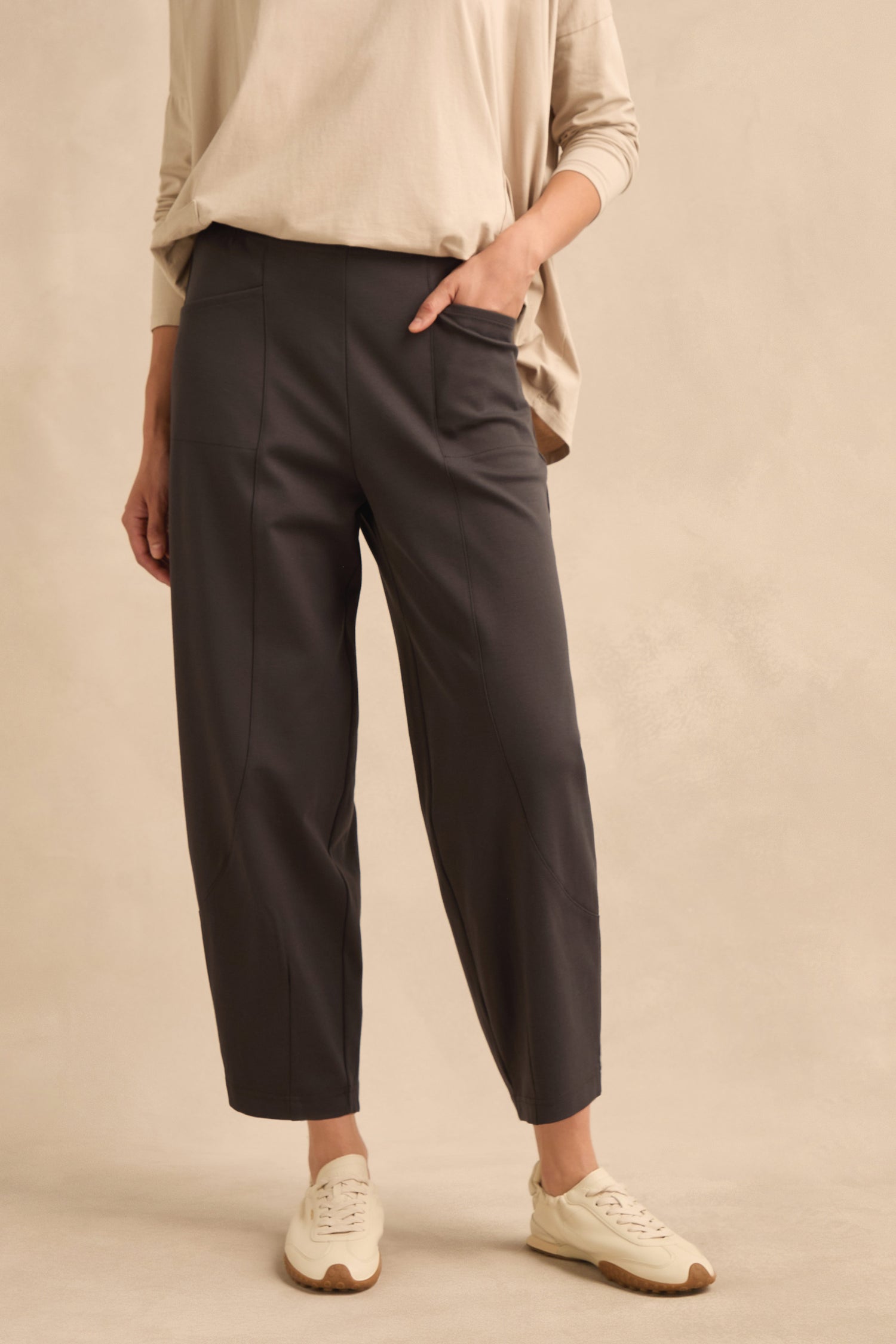 A person wearing a loose tan top, Slate Ponte Bubble Trousers with an elasticated waist, and white sneakers stands against a beige background with one hand in a pocket.