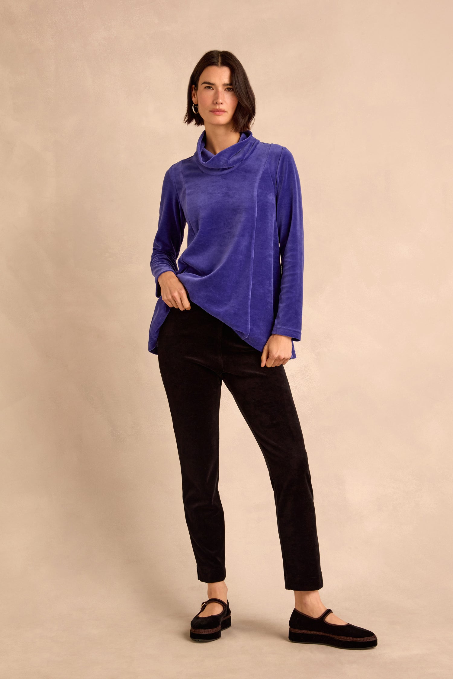 A woman stands against a beige background, wearing a blue long-sleeve cowl neck top, Black Velvet Jersey Slim Trousers, and black shoes.