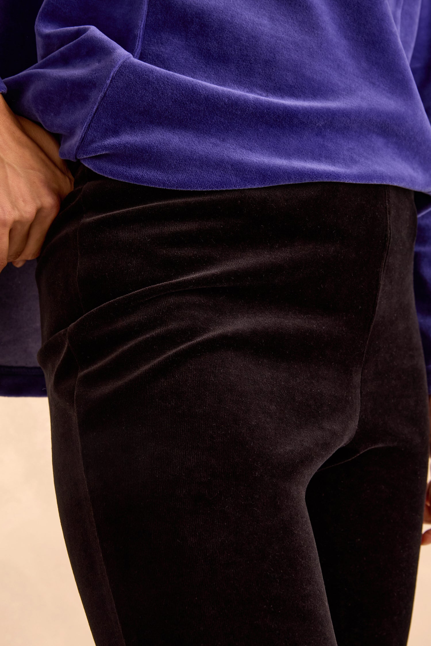 A person wears Black Velvet Jersey Slim Trousers paired with a blue long-sleeve top, showcasing a slim silhouette and posing with one hand on their hip.