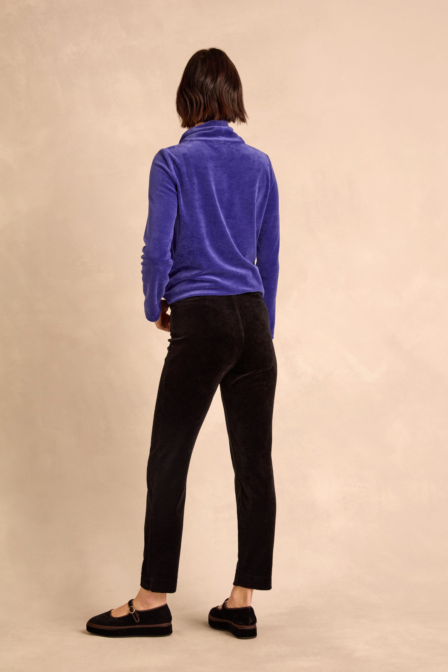 A person stands facing away from the camera against a beige background, wearing a blue long-sleeve top, Black Velvet Jersey Slim Trousers, and black slip-on shoes.