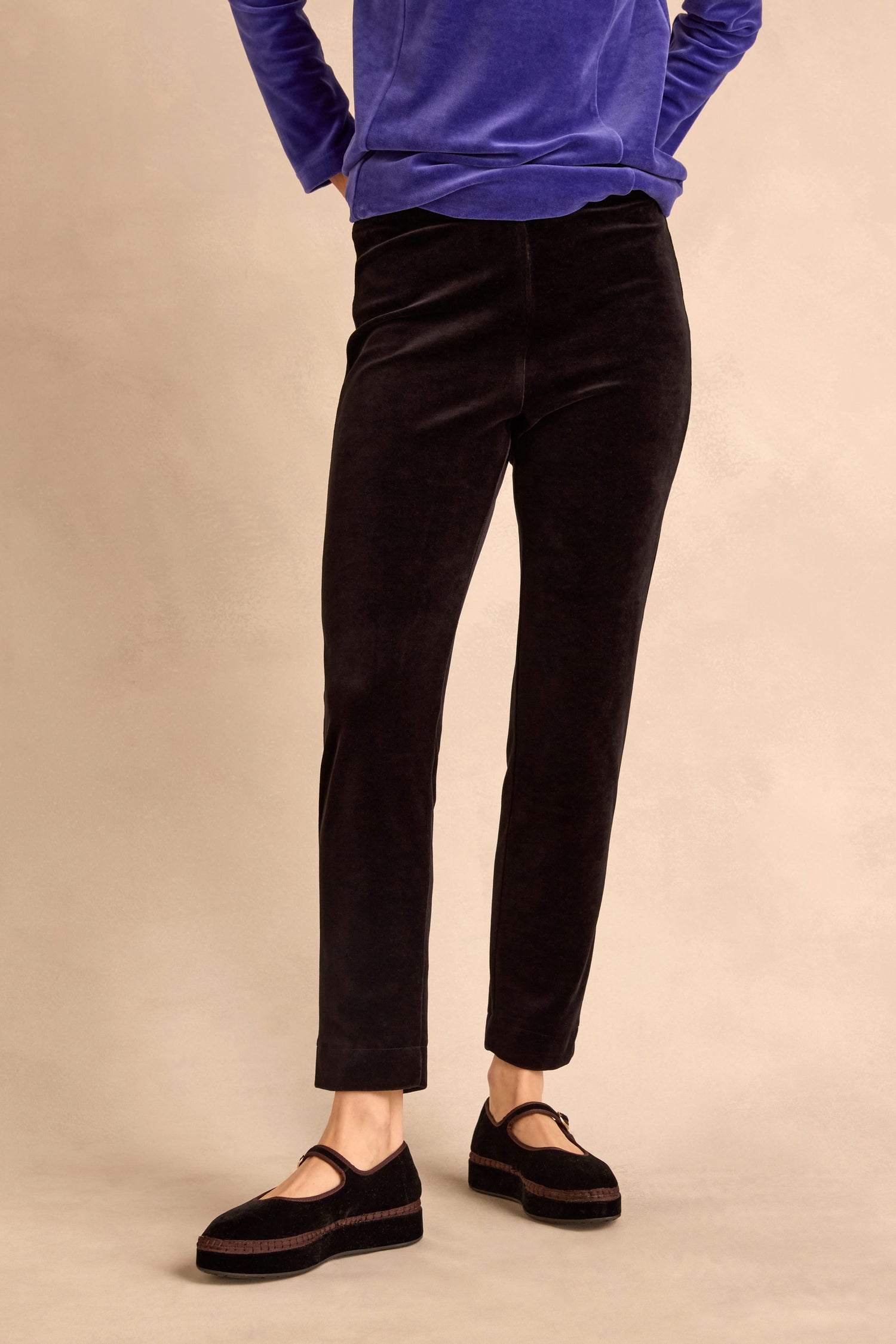 Wearing the Black Velvet Jersey Slim Trousers for a slim silhouette, paired with black Mary Jane shoes and a long-sleeved blue top, this person stands against a neutral background with hands in their pockets.