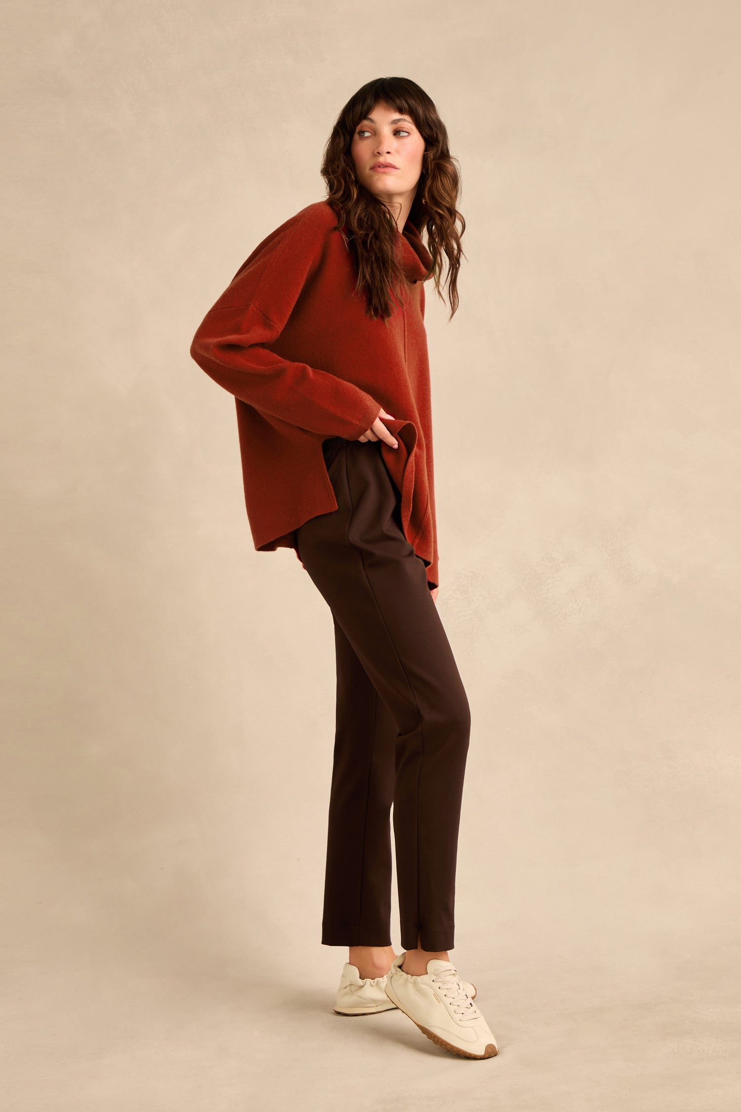 A woman stands against a beige background wearing a rust-colored sweater, the Chocolate Ponte Slim Leg Trousers with a pull-on elasticated waist, and white sneakers.
