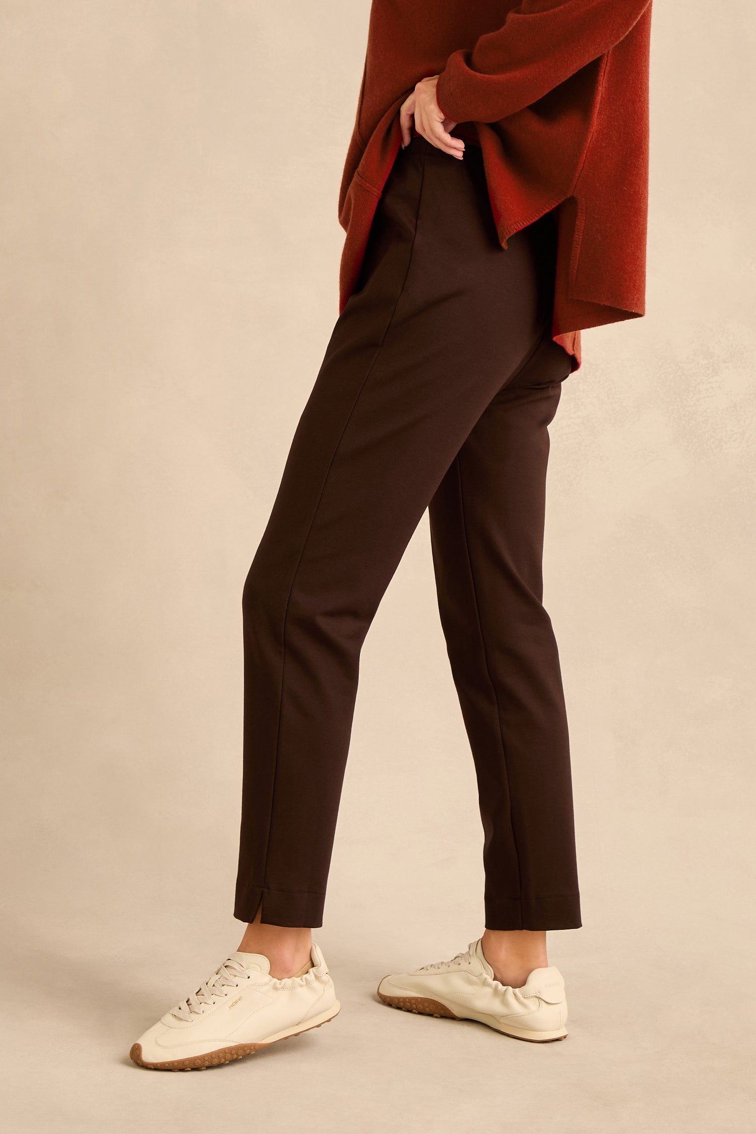 A person wearing an oversized red sweater, the Chocolate Ponte Slim Leg Trousers featuring a pull-on elasticated waist in chocolate brown, and white sneakers stands against a beige background. Only the lower half of the body is visible.