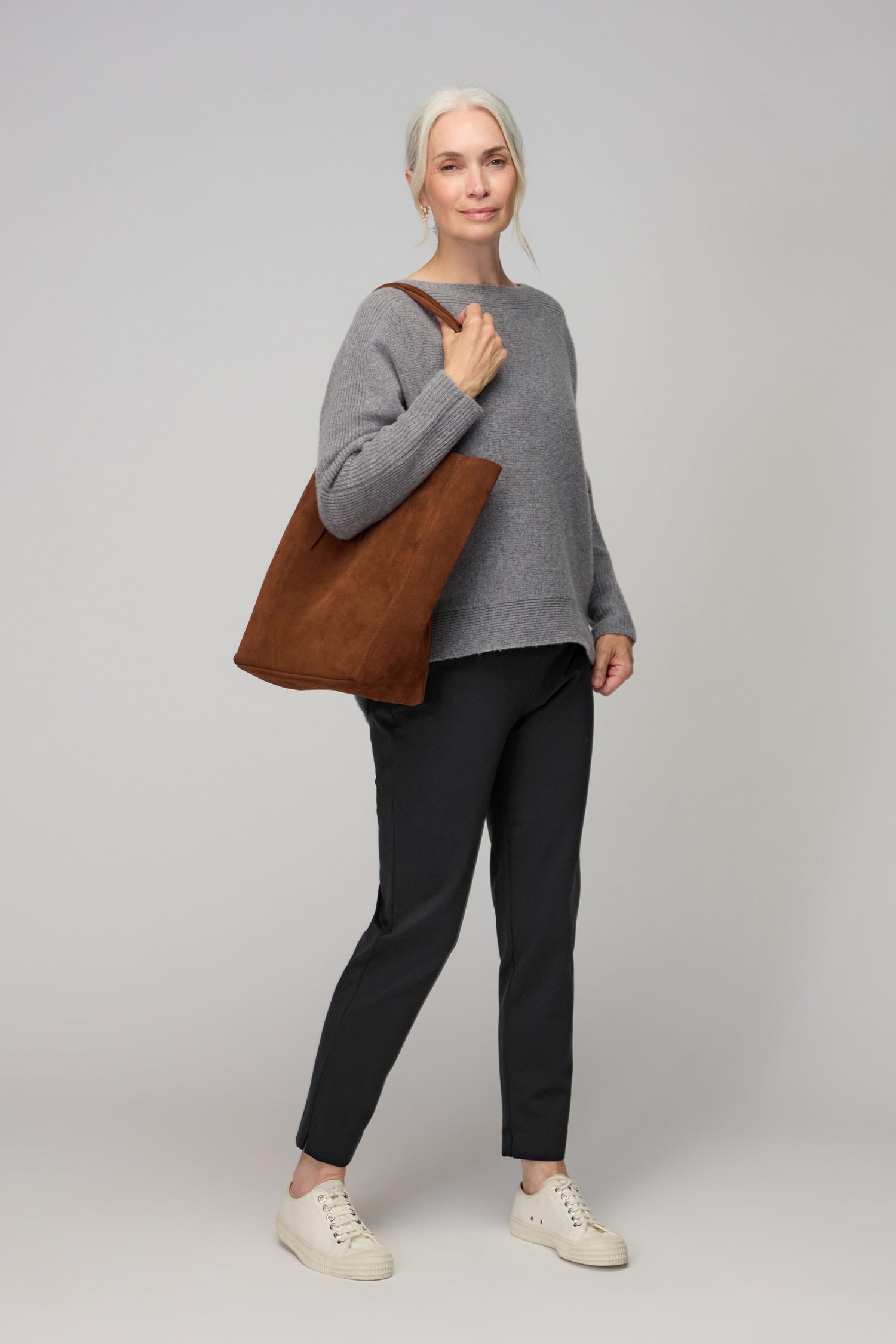 An older woman with gray hair in a ponytail, wearing a gray sweater, Ponte Slim Leg Trousers, white sneakers, and carrying a brown tote bag stands against a plain light background.