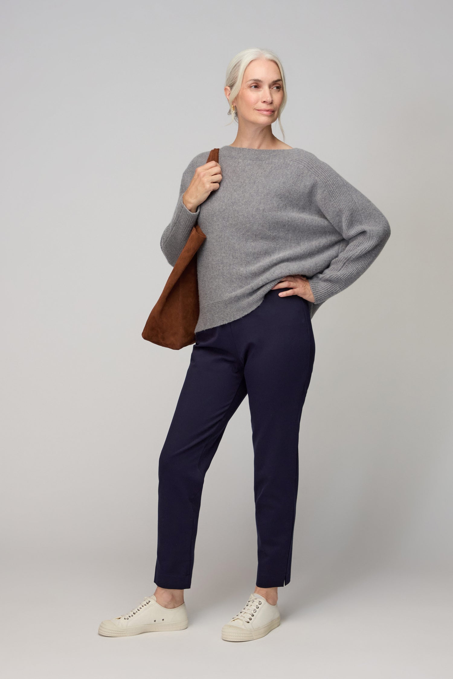 A woman stands against a plain background wearing a gray sweater, white sneakers, a brown shoulder bag, and Ponte Slim Leg Trousers—highlighting these chic trousers as versatile wardrobe essentials.