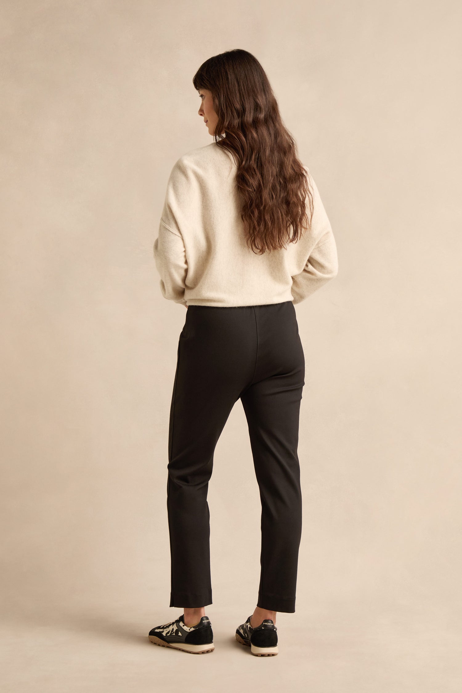 A person with long brown hair stands facing away, wearing a cream sweater, Black Ponte Slim Leg Trouser, and black sneakers against a beige background.