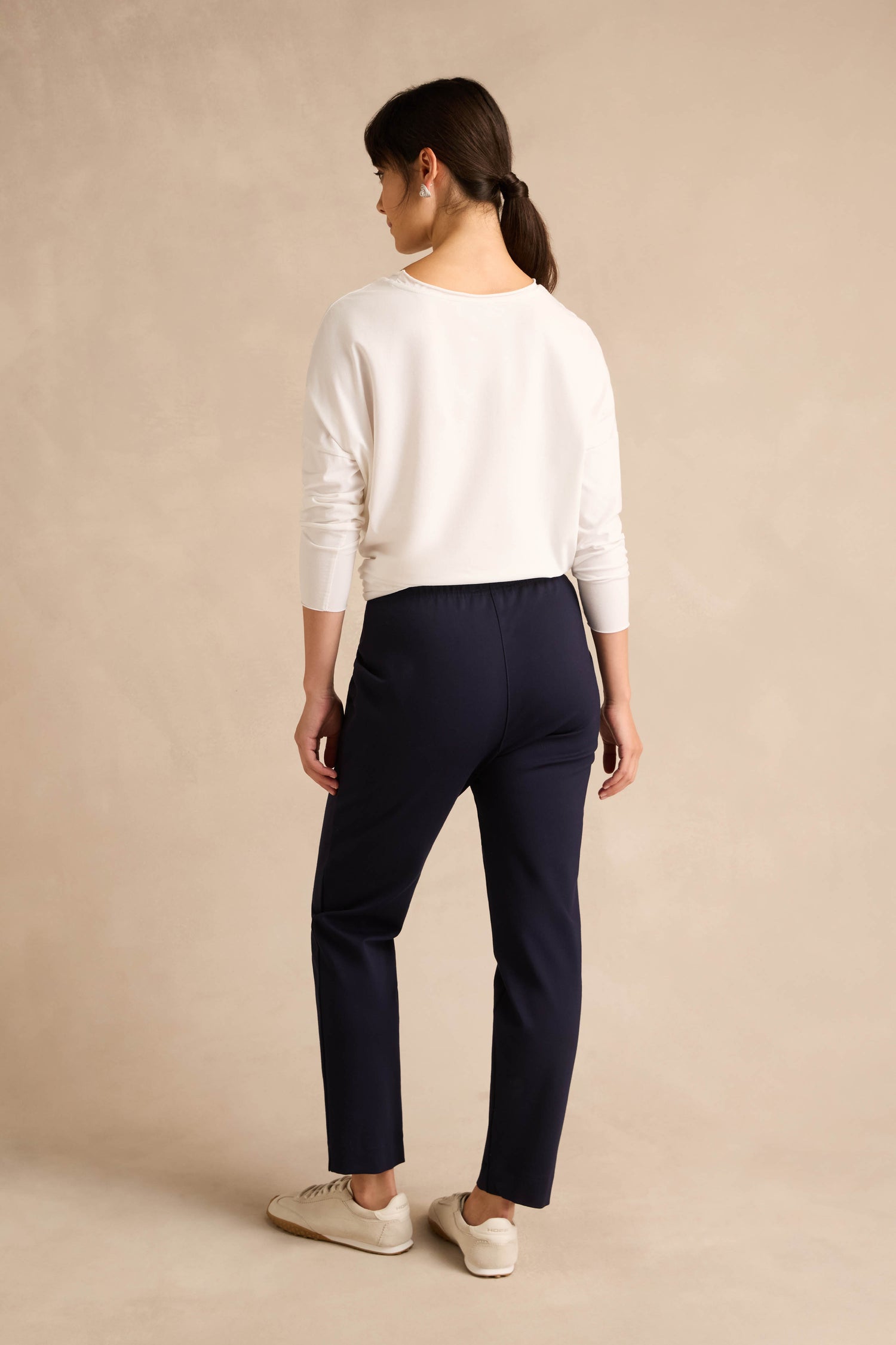 A woman stands with her back to the camera, wearing a white long-sleeve shirt, beige shoes, and the Midnight Ponte Slim Leg Trouser in navy against a plain beige background.