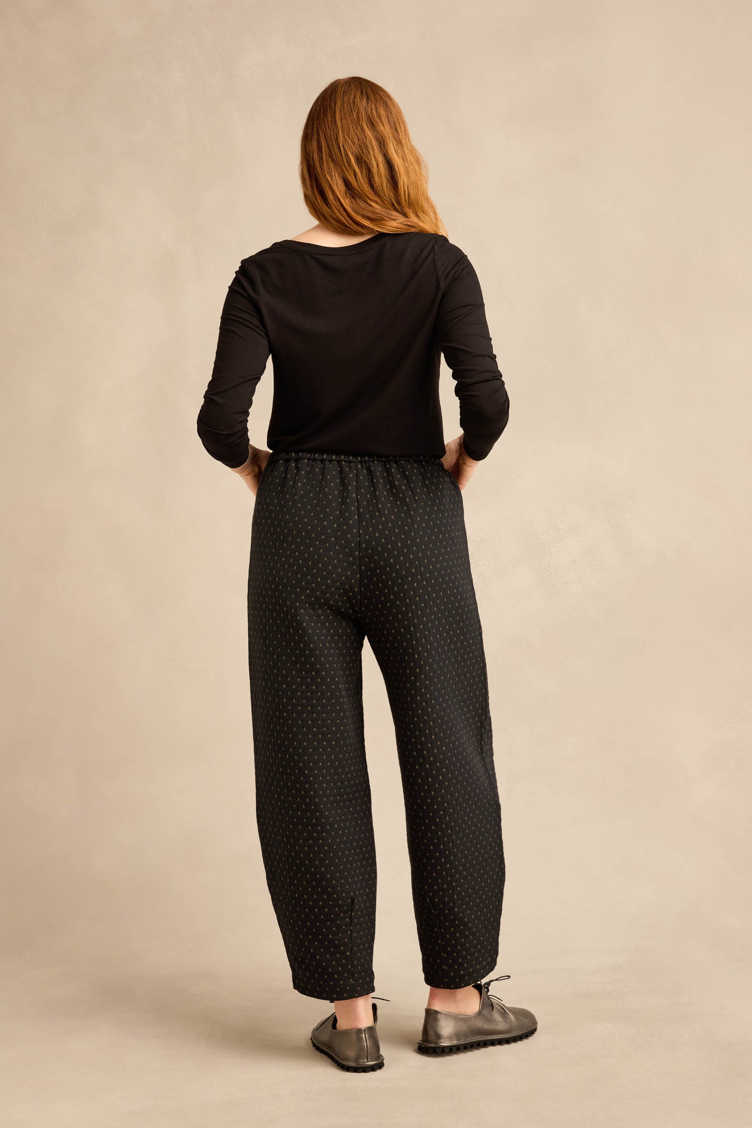 A woman stands with her back to the camera, wearing a black long-sleeve shirt, black shoes, and Square Jacquard Bubble Trousers in a cropped bubble leg style against a neutral background.