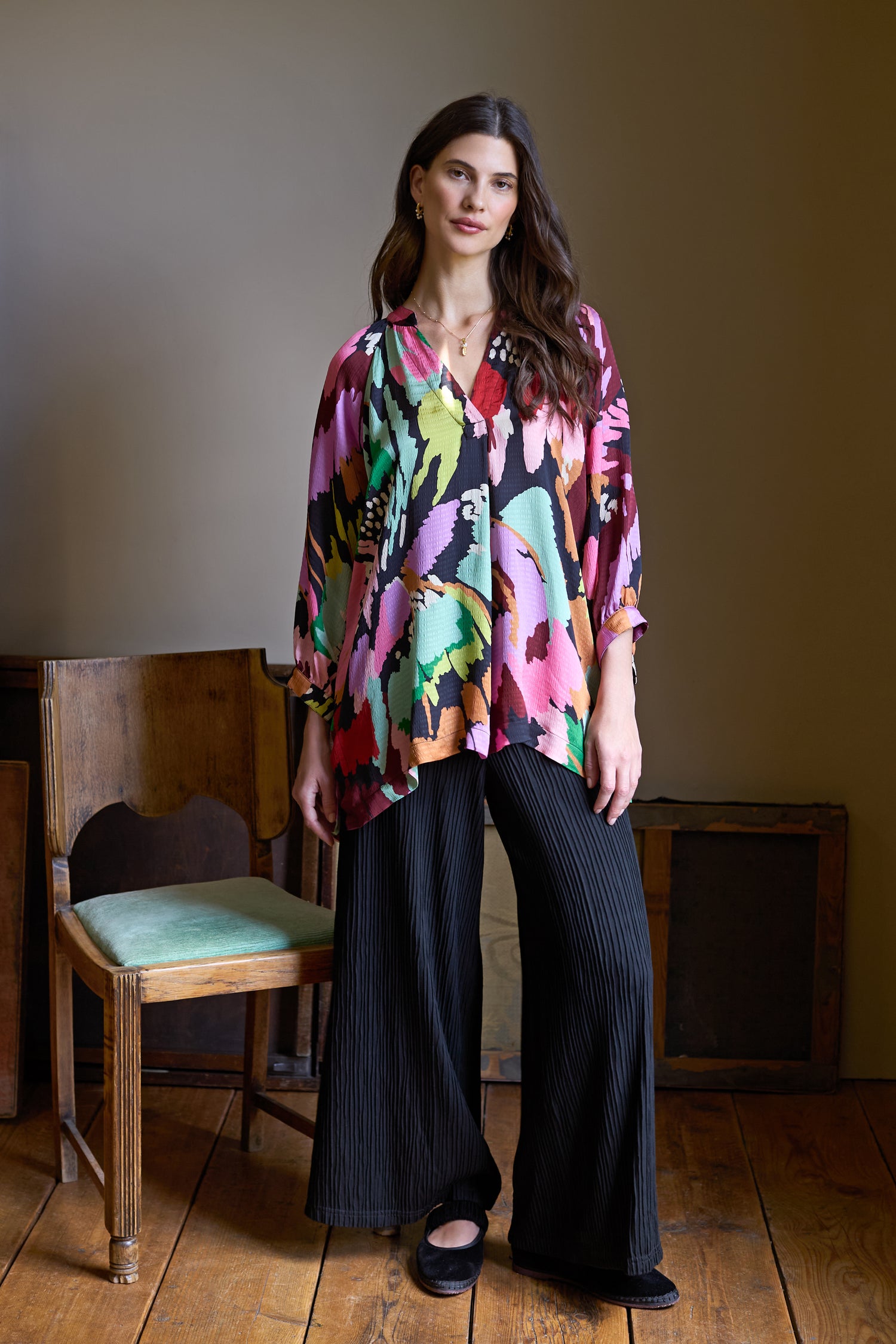 A woman stands indoors on wooden floors wearing a colorful patterned blouse, the Textured Wave Jersey Wide Trousers (SPP6544-TWVJ), and black shoes. Two wooden chairs and framed objects are visible in the background.