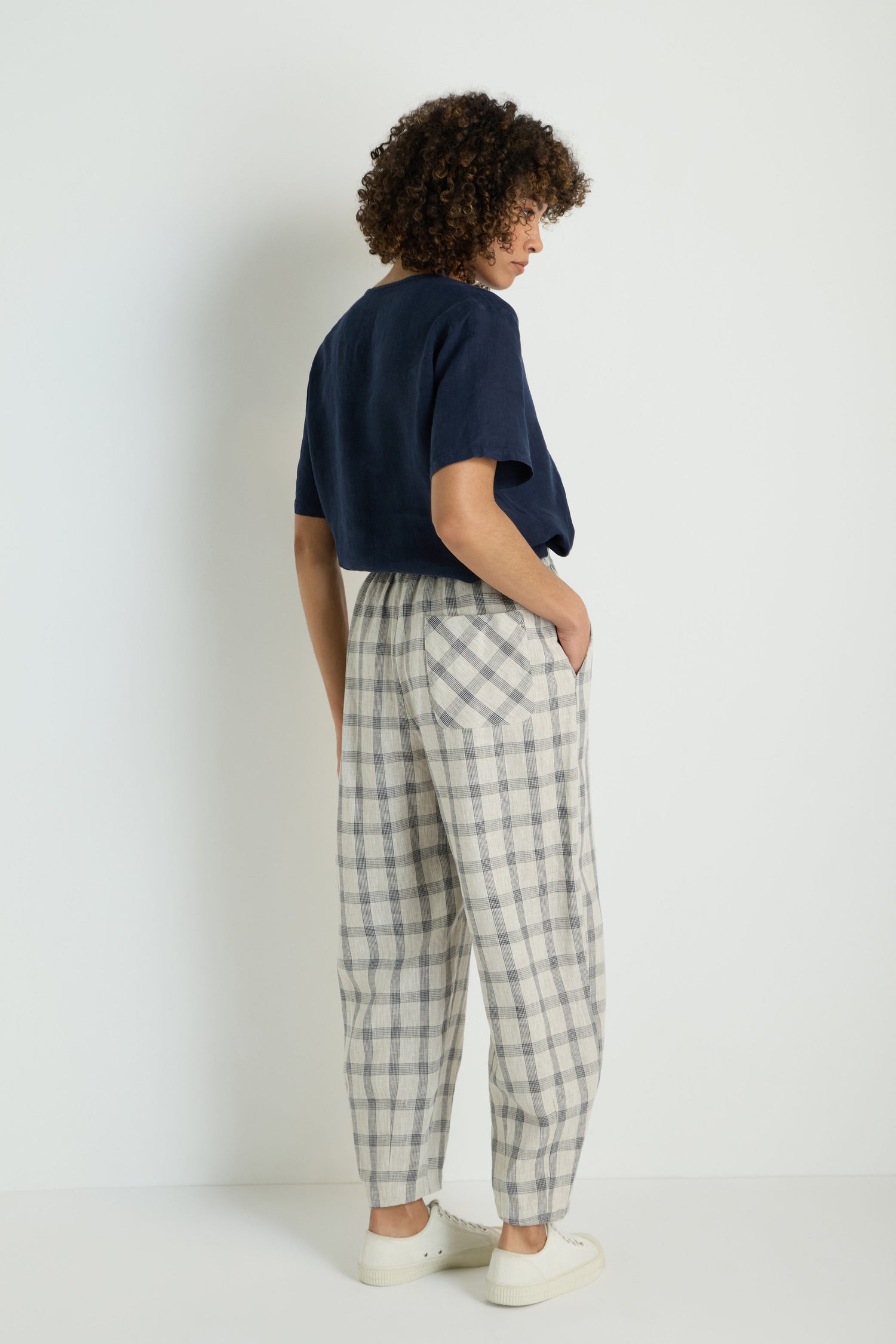 A woman stands against a white wall, wearing a short-sleeve cream top, Grid Check Linen Bubble Trouser featuring its signature bubble shape, and brown slippers.