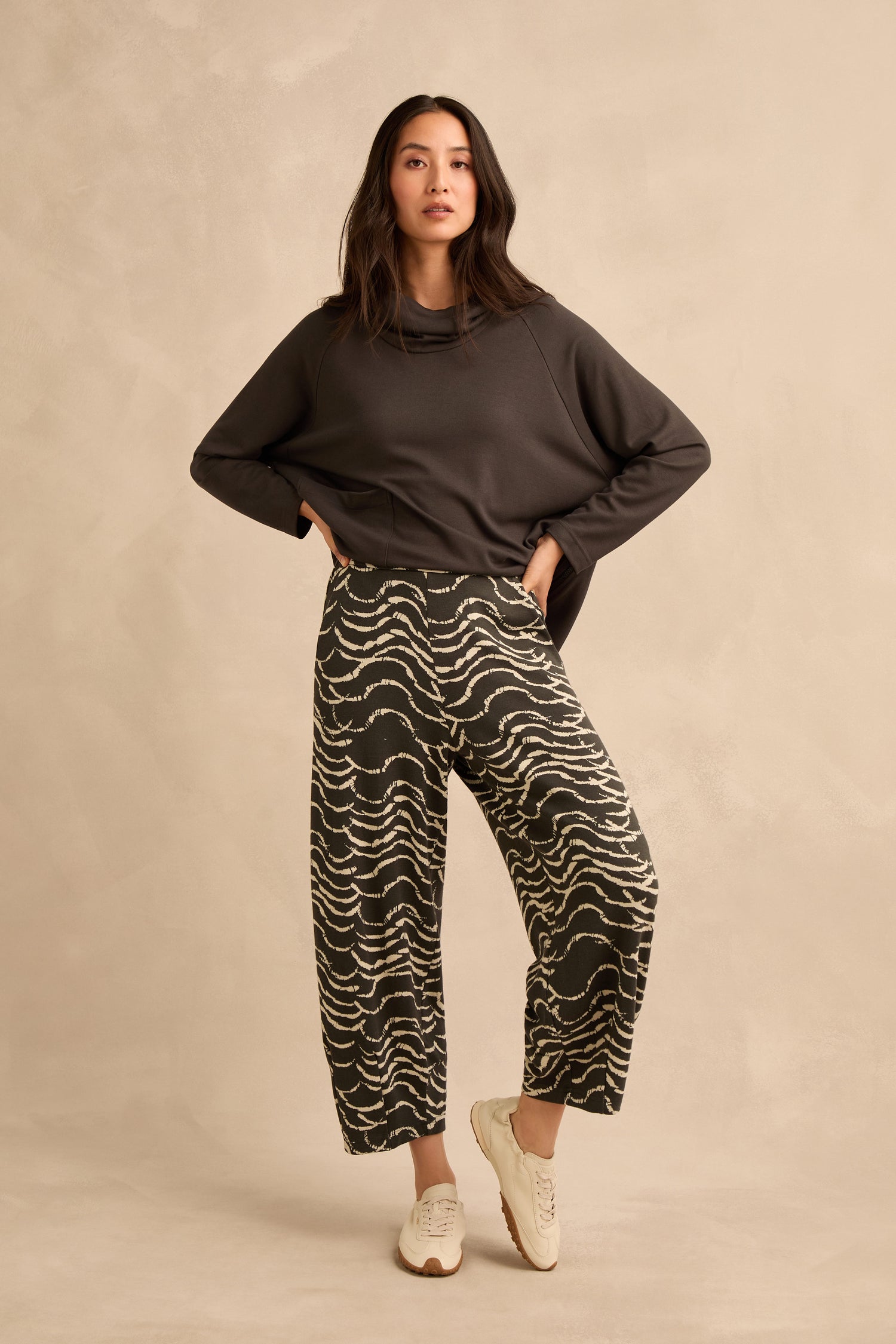 A woman stands against a beige background wearing a dark long-sleeve top, white sneakers, and the Dancing Waves Jersey Bubble Trousers, featuring a stretch jacquard wide-leg print. She looks directly at the camera with one hand on her hip.