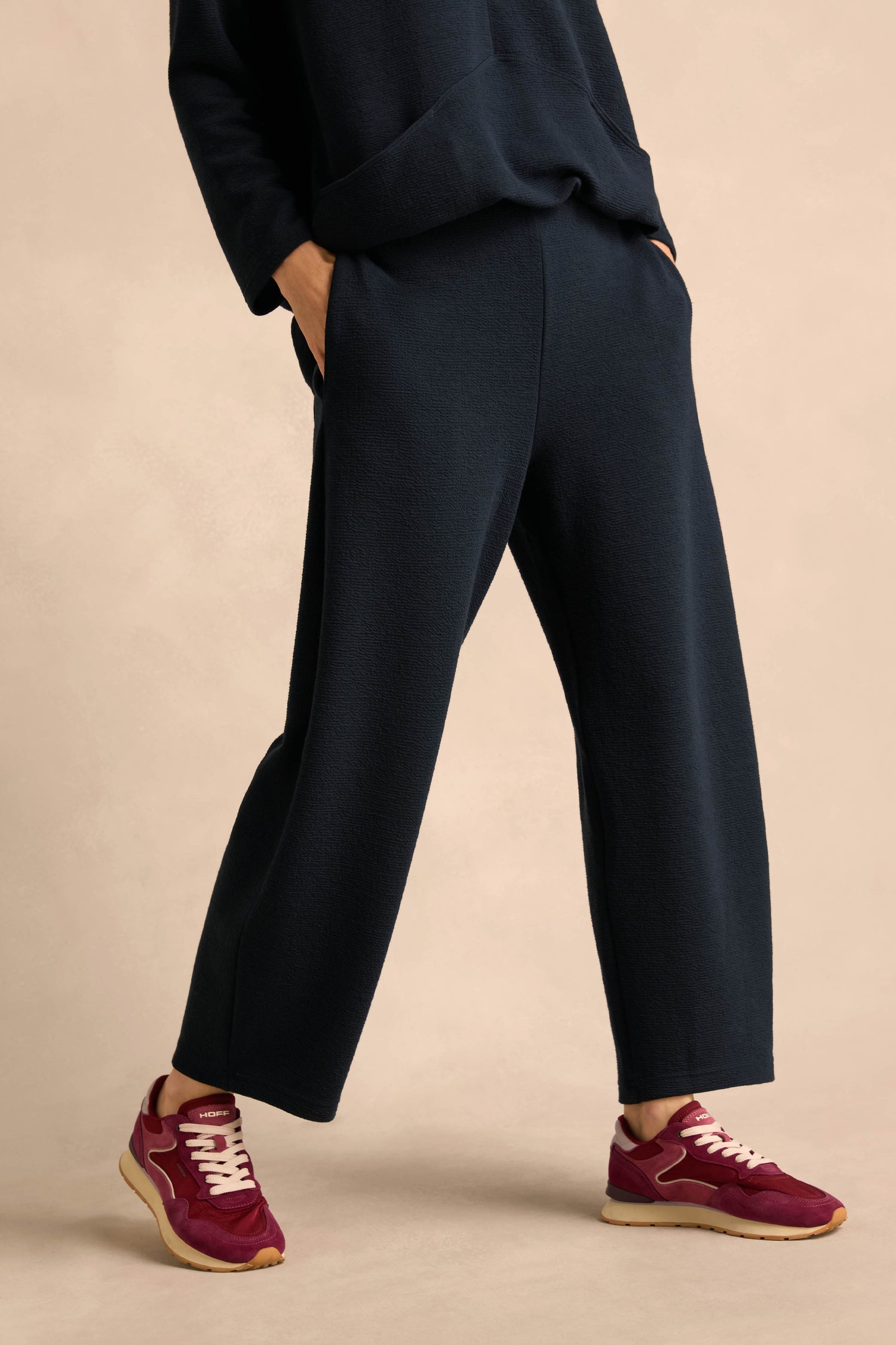 A person stands against a neutral background wearing Double Jersey Crinkle Trousers in navy blue with a matching top, hands in pockets, and paired with burgundy sneakers. These relaxed-leg Double Jersey Crinkle Trousers are made in England, offering both comfort and style.