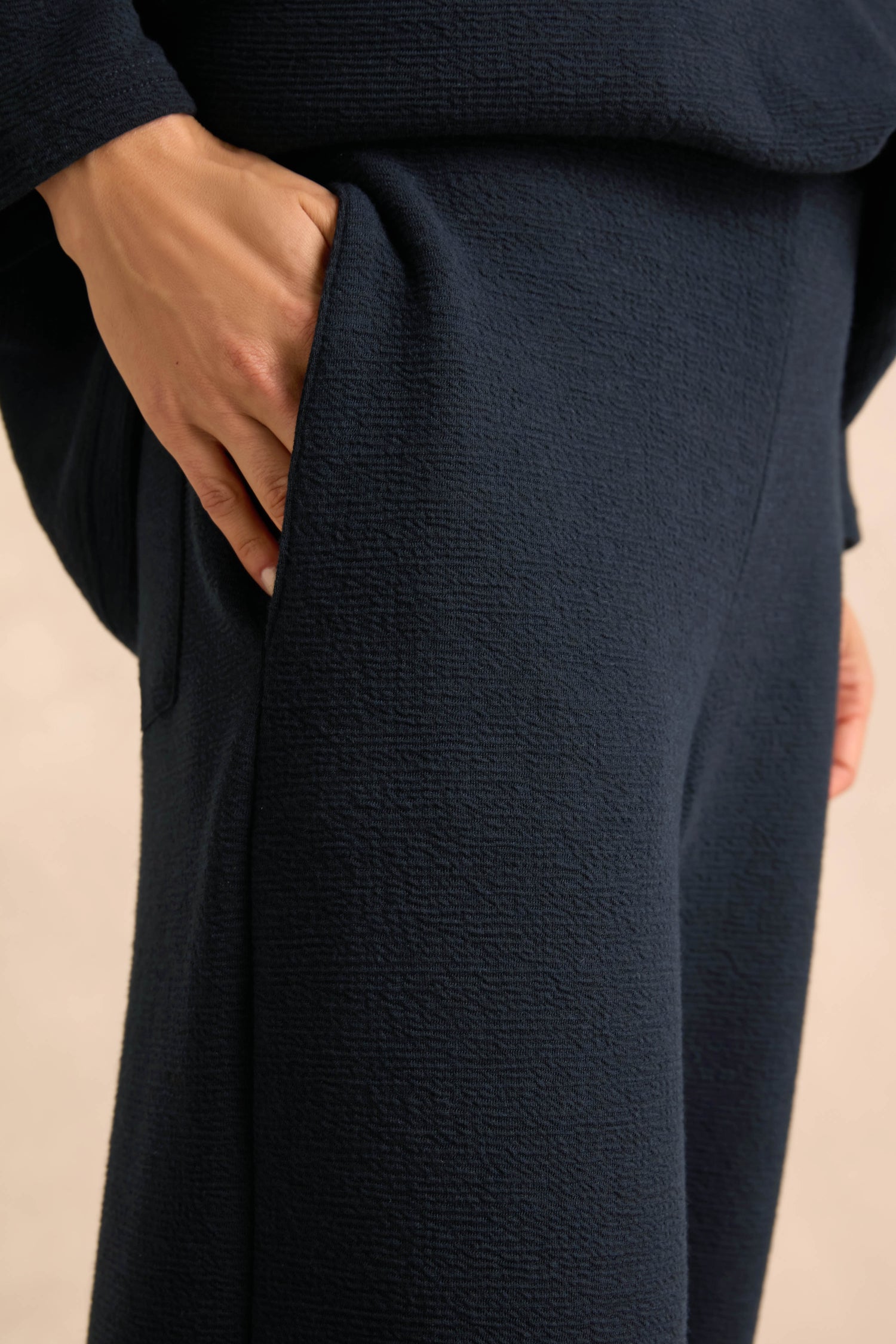 A close-up shows a hand resting in the pocket of the Double Jersey Crinkle Trousers in dark blue, with part of the wearer’s arm and hip visible. Made in England, these relaxed-leg trousers highlight distinctive textured fabric and expert craftsmanship.