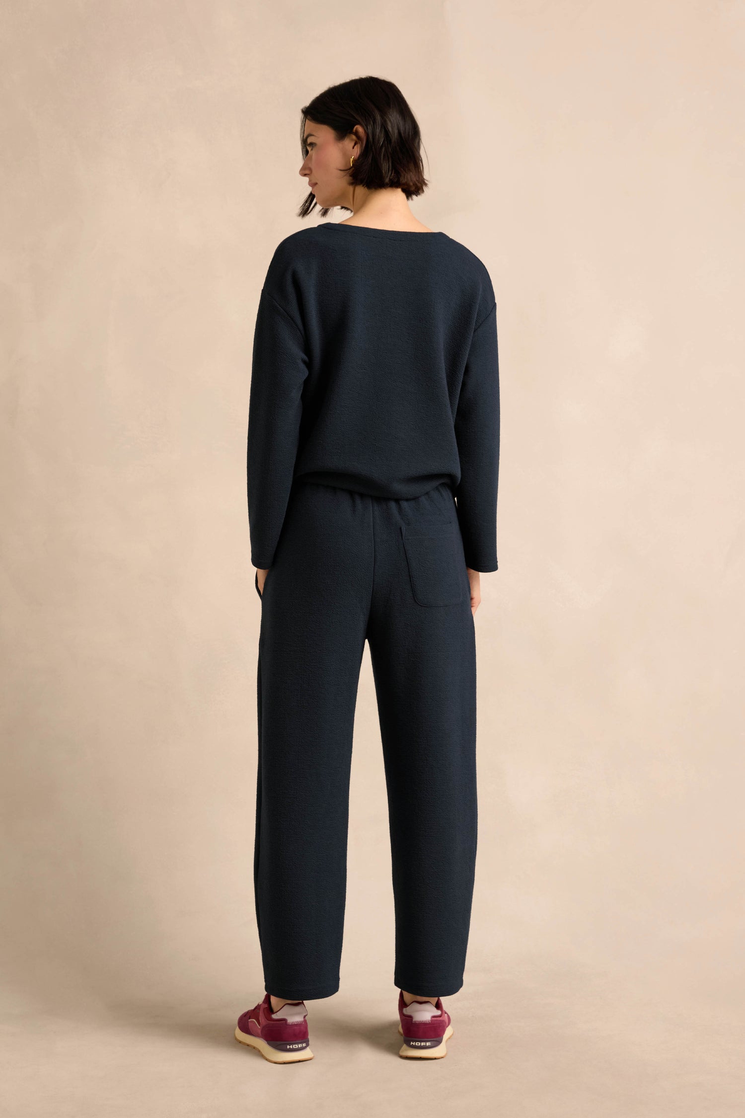 A person stands facing away, dressed in a dark long-sleeve top and the Double Jersey Crinkle Trousers—relaxed-leg trousers crafted in England—paired with maroon sneakers, set against a neutral background.