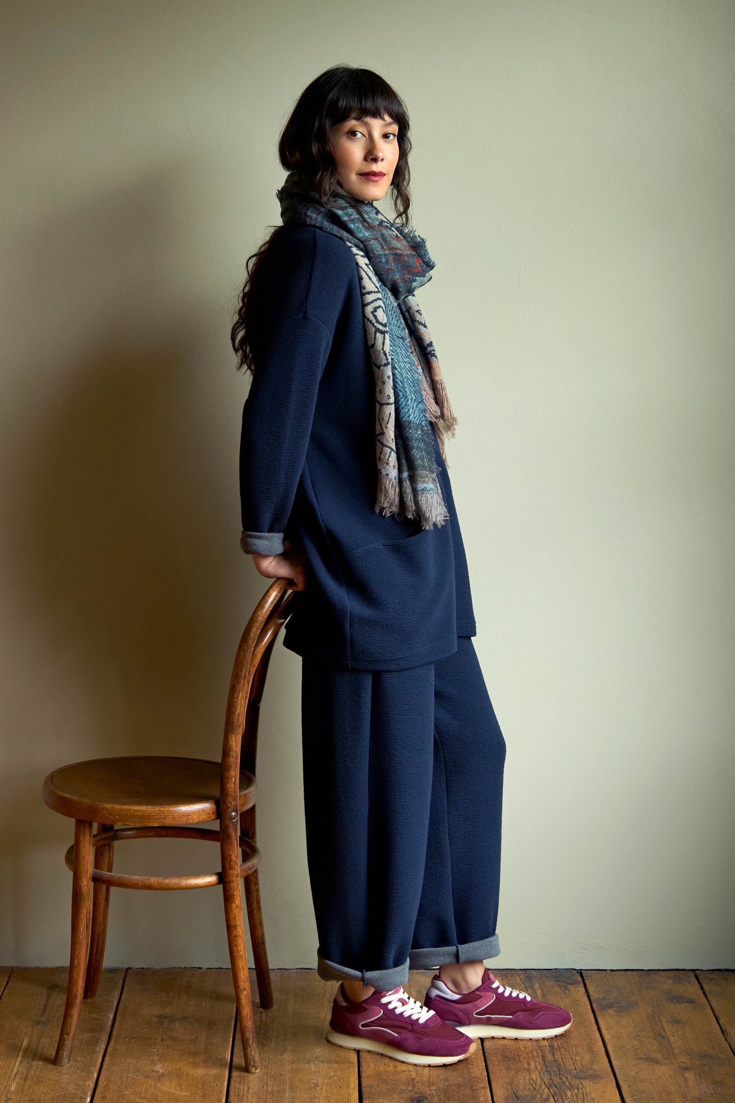 A woman in a dark blue top and matching Double Jersey Crinkle Trousers stands leaning against a wooden chair, accessorized with a patterned scarf and burgundy sneakers.