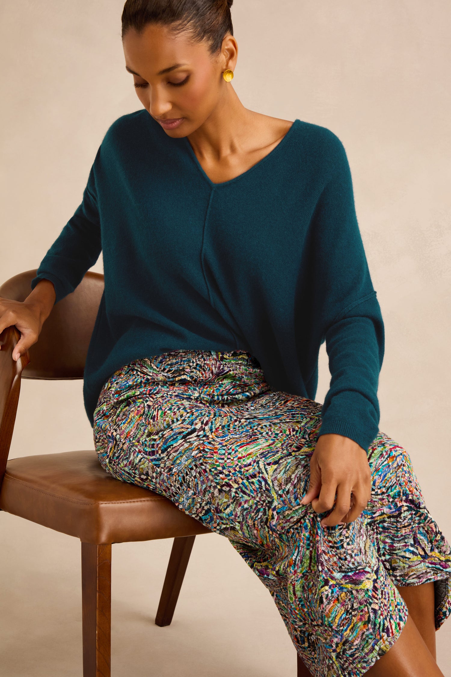 Woman seated on a brown chair wearing a teal V-neck sweater, the Patchwork Fields Jersey Tube Skirt with multicolored prints, and gold earrings, looking down with one hand resting on her knee.