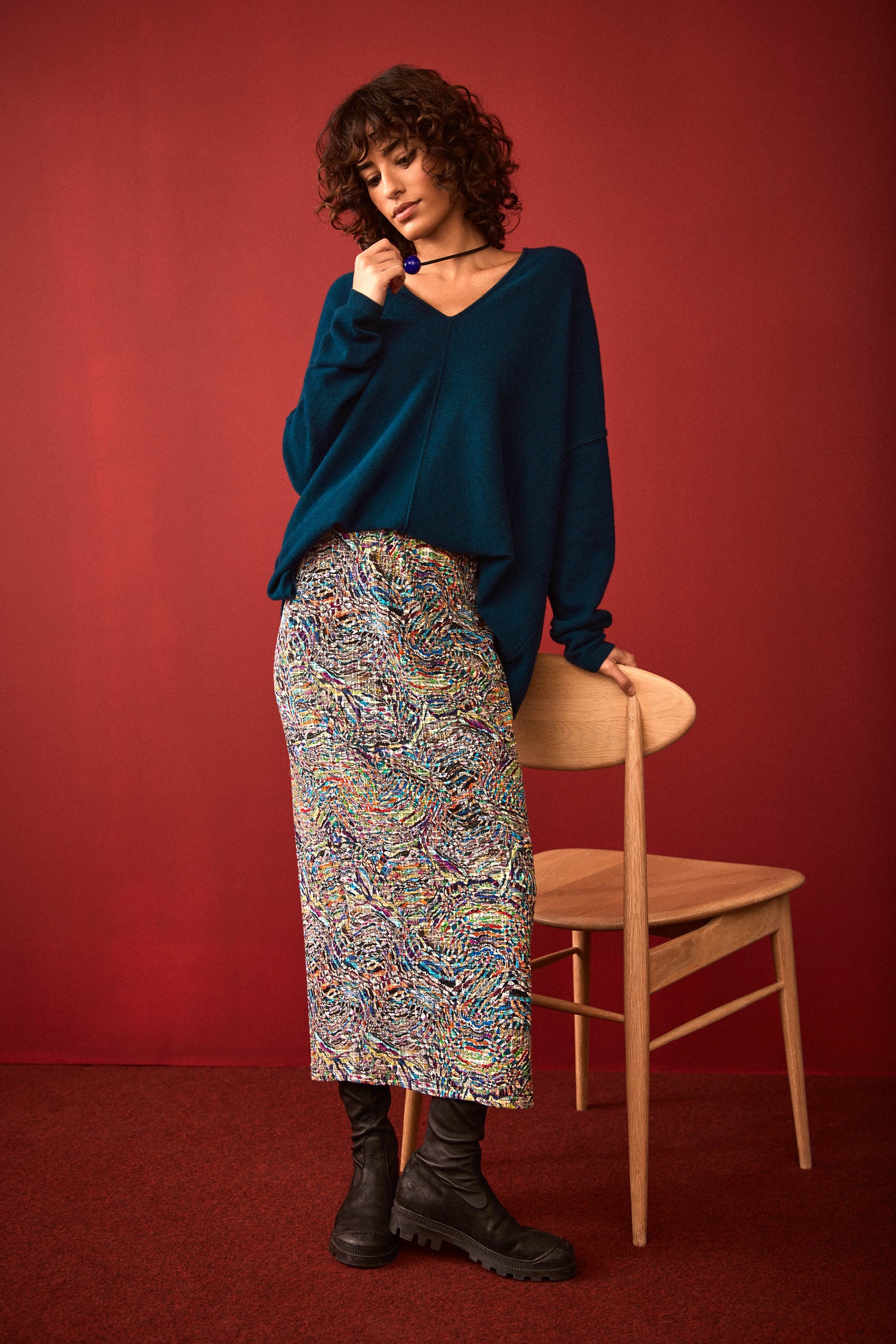 A woman stands in front of a red wall, wearing a teal sweater and the Textured Prism Jersey Tube Skirt—a multicolored patterned midi tube skirt with an elasticated waist—paired with black boots. One hand rests on a wooden chair.