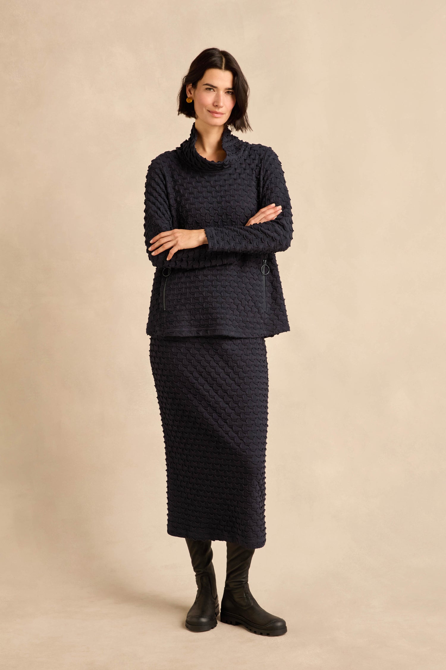 A woman stands against a beige background, wearing a textured stretch jersey dark navy long-sleeve top and the Pocket Jersey Tube Skirt in matching navy, paired with black boots.