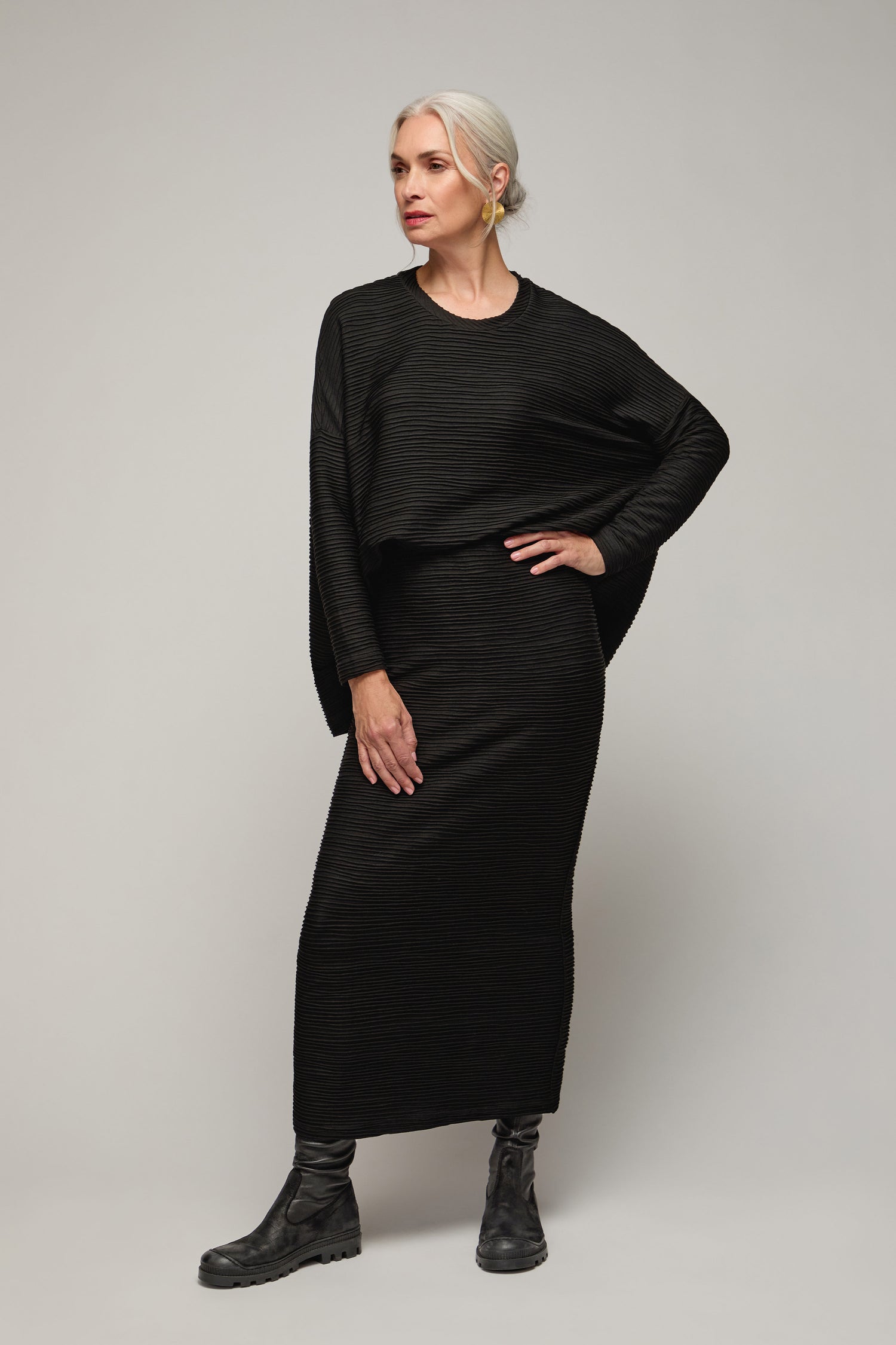 A woman with white hair poses in a black Textured Wave Jersey Tube Skirt and matching long-sleeve top, paired with black boots, standing against a plain light background.