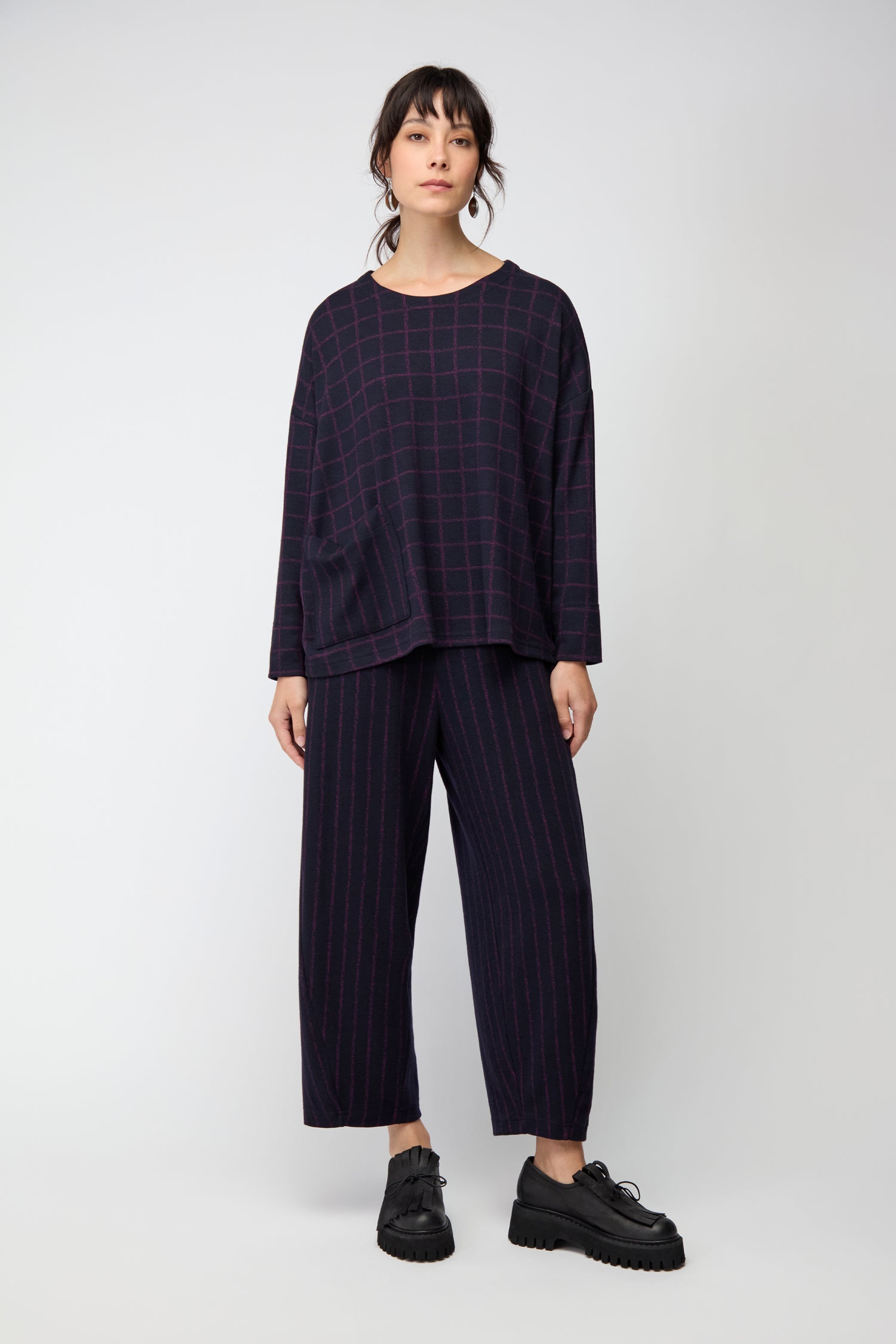 A woman stands against a plain background wearing the Small Broken Check & Stripe Boxy Top with matching pants, styled with black platform shoes and hoop earrings.