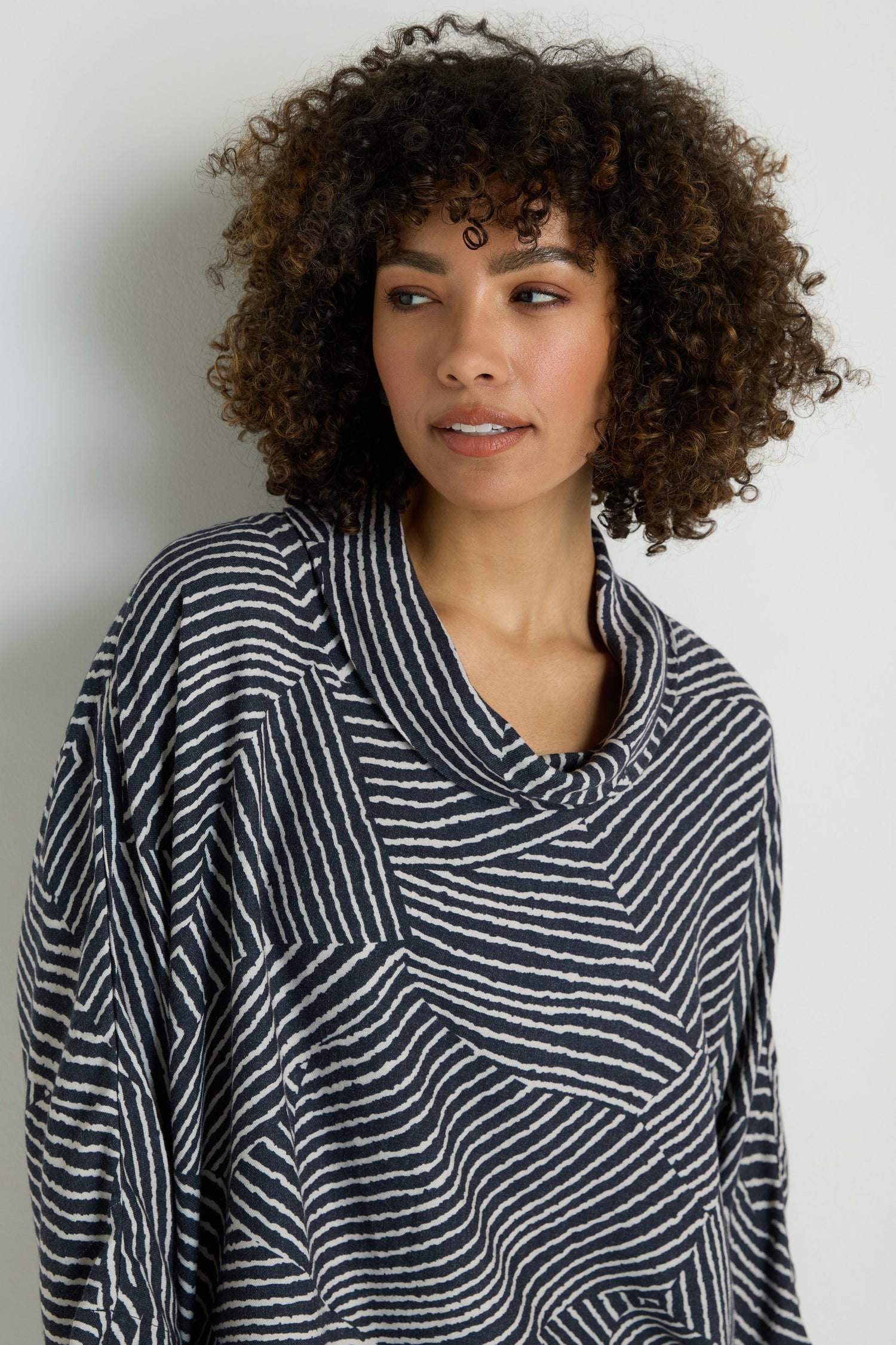 A woman with long blonde hair wears the Linear Prism Lines Cowl Neck Top in navy and white geometric print, made from soft brushed viscose, standing against a plain light background and looking down with a neutral expression.